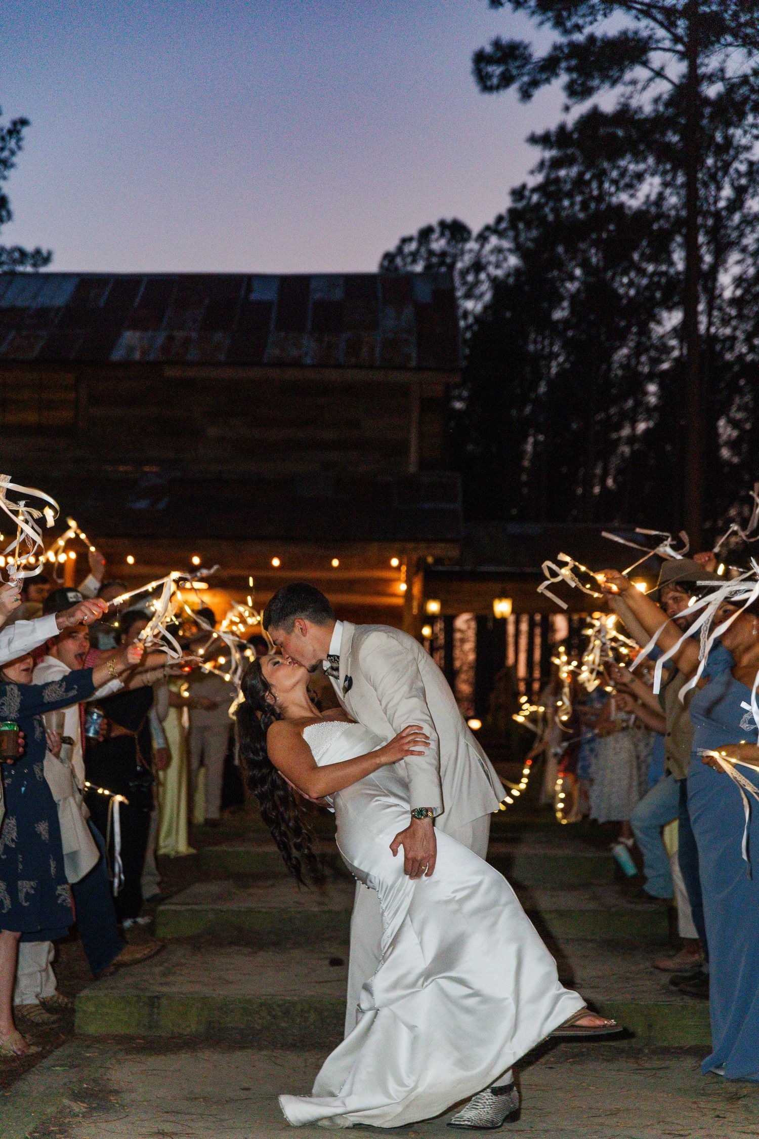 Groom dips bride for a kiss during sparkler exit at dusk outside a rustic waterfront wedding venue.