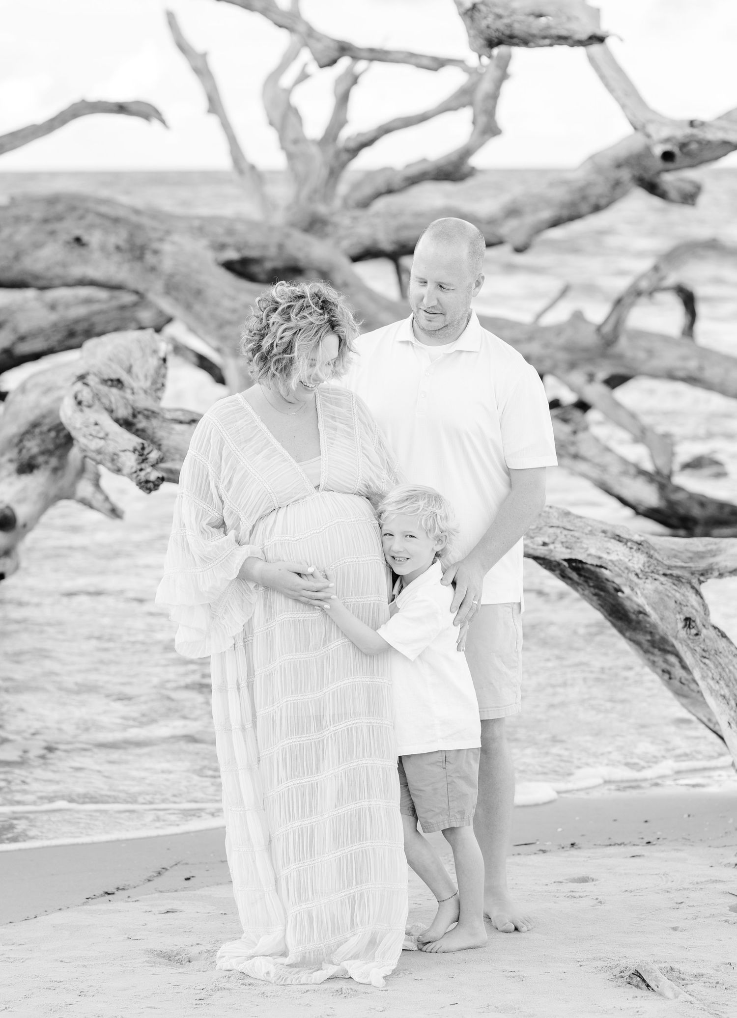 A black and white beach photoshoot features a family posing among driftwood on the sand.