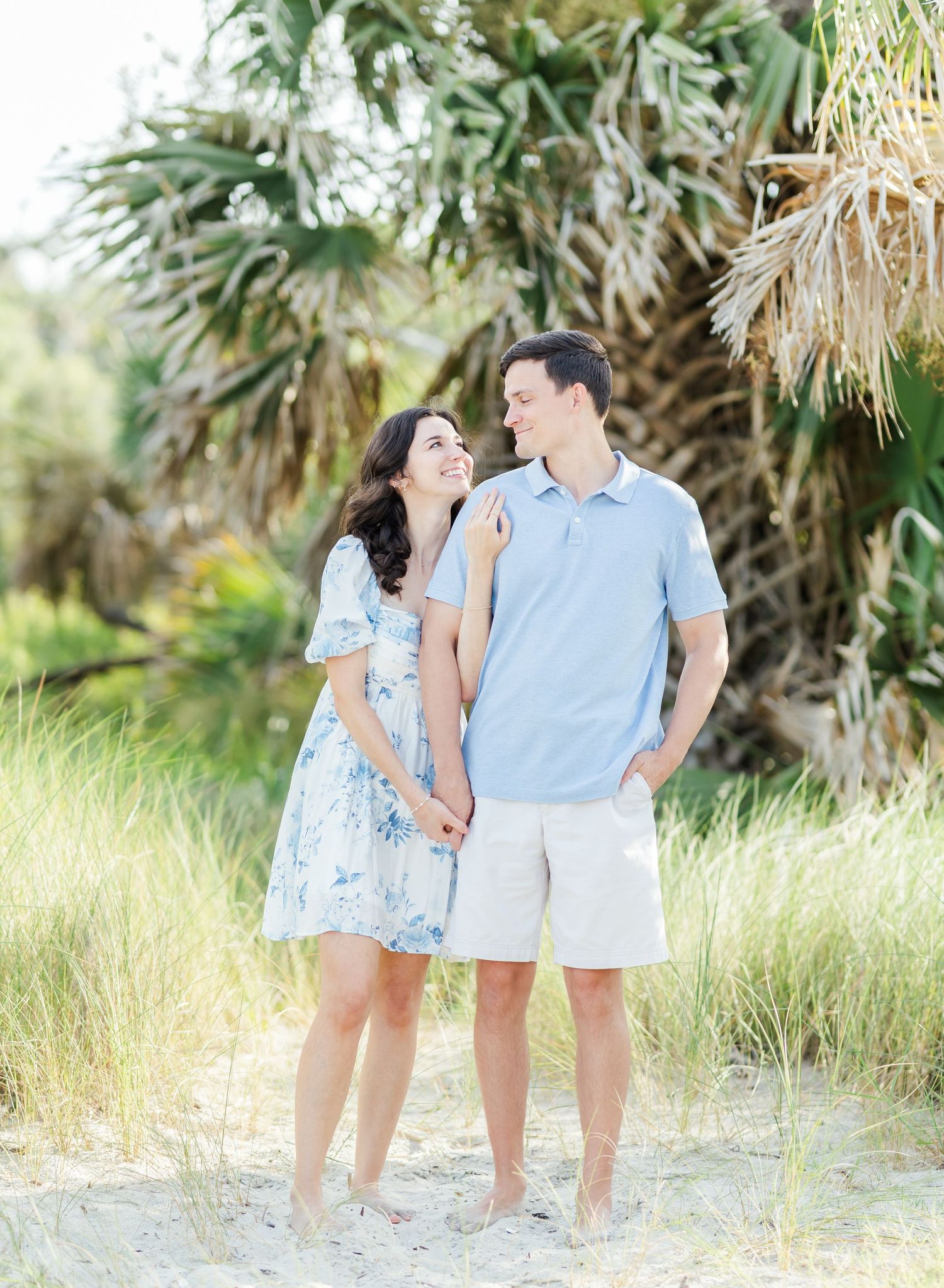 Romantic couple laughs together while walking along a sandy coastal path surrounded by palm trees.