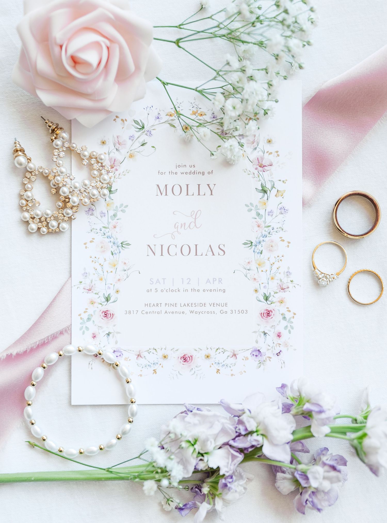 Wedding invitation with blush pink roses, rings, pearl jewelry and delicate floral details arranged on white background.