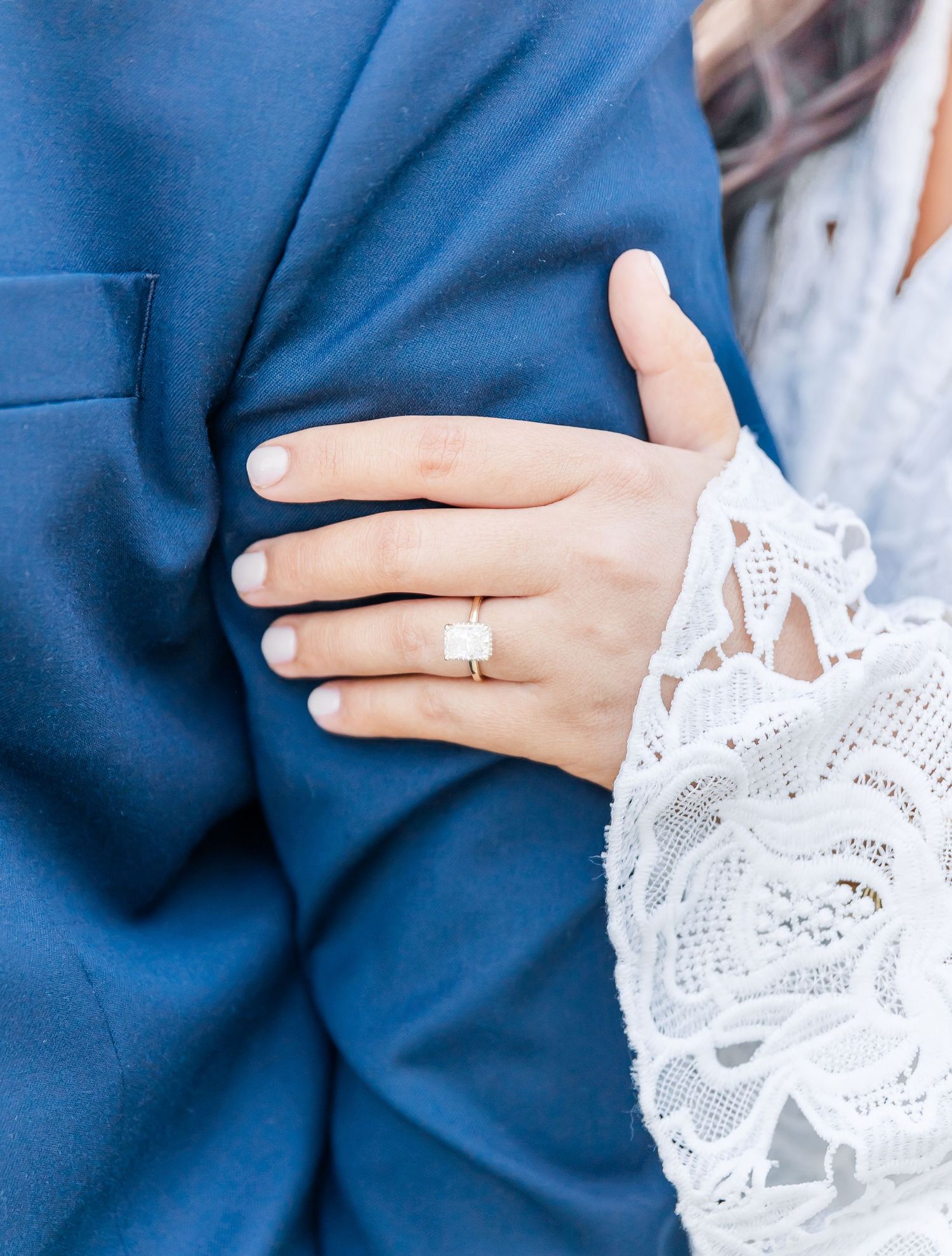 A delicate white lace sleeve touches a navy blue suit jacket, showcasing an elegant diamond engagement ring.