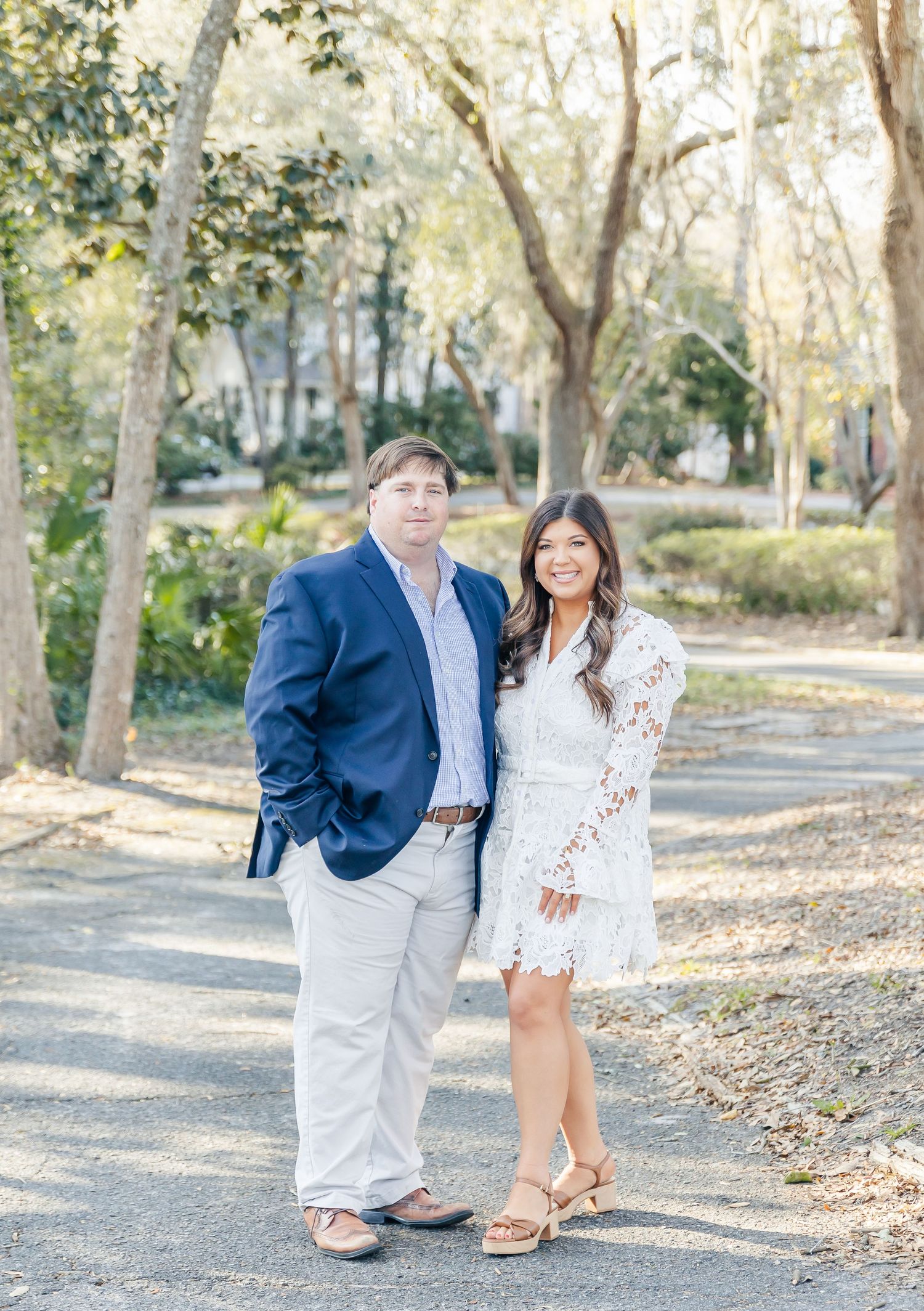 A couple poses together on a sunlit walking path surrounded by trees in a romantic outdoor setting.