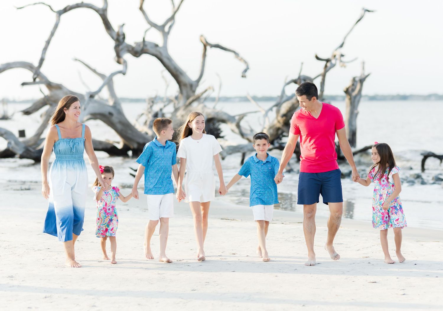 A group walks together along a pristine white beach with driftwood and calm waters in the background.
