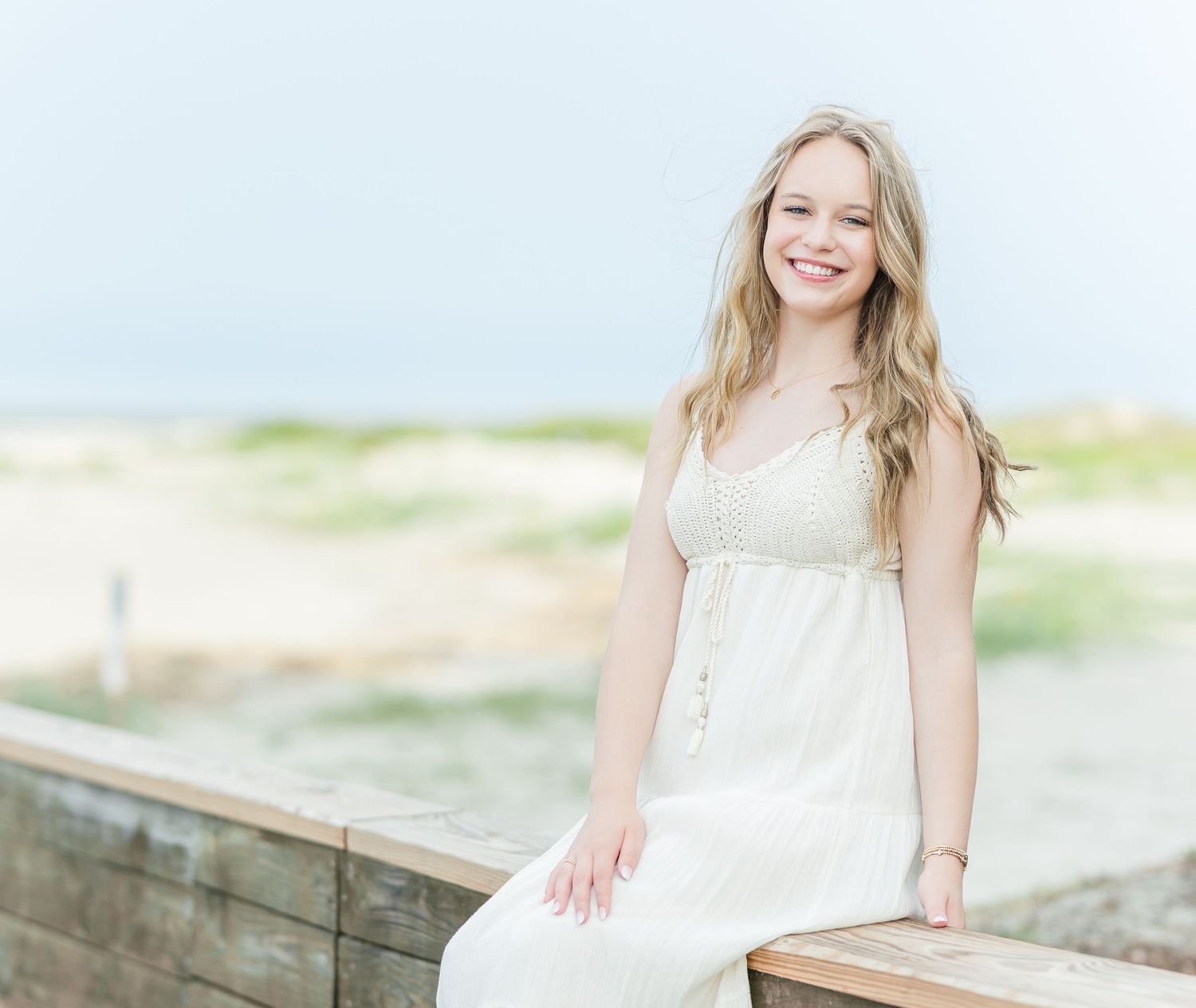 Someone in a white summer dress sits on a weathered wooden boardwalk by the beach on a bright sunny day.