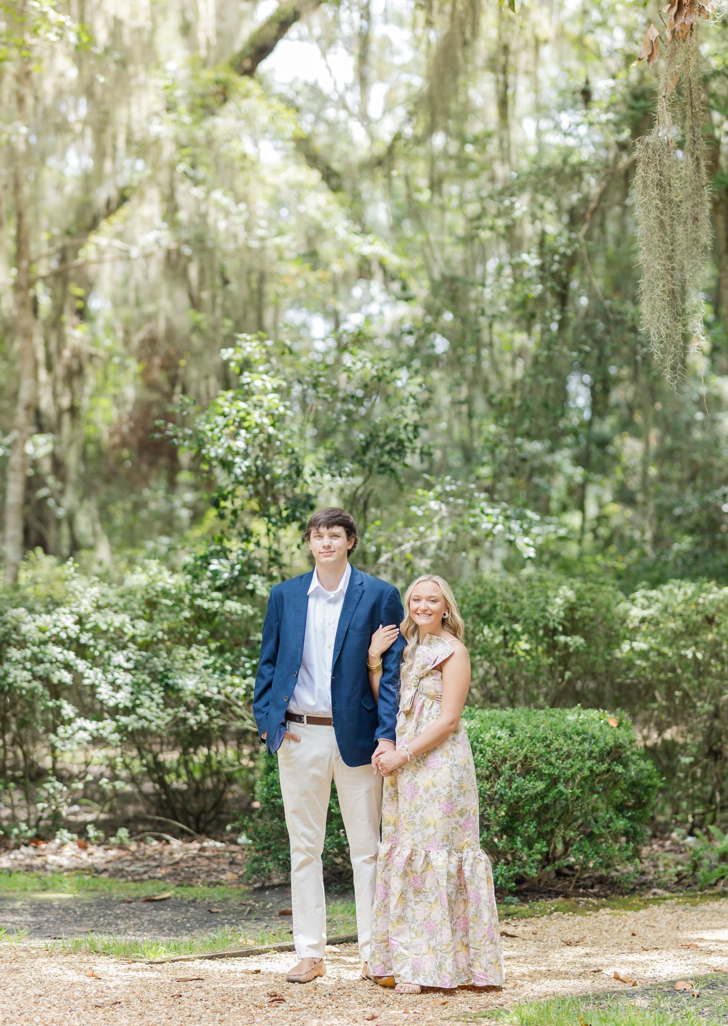 Romantic couple poses on a dirt path under Spanish moss-draped trees in a dreamy garden setting.