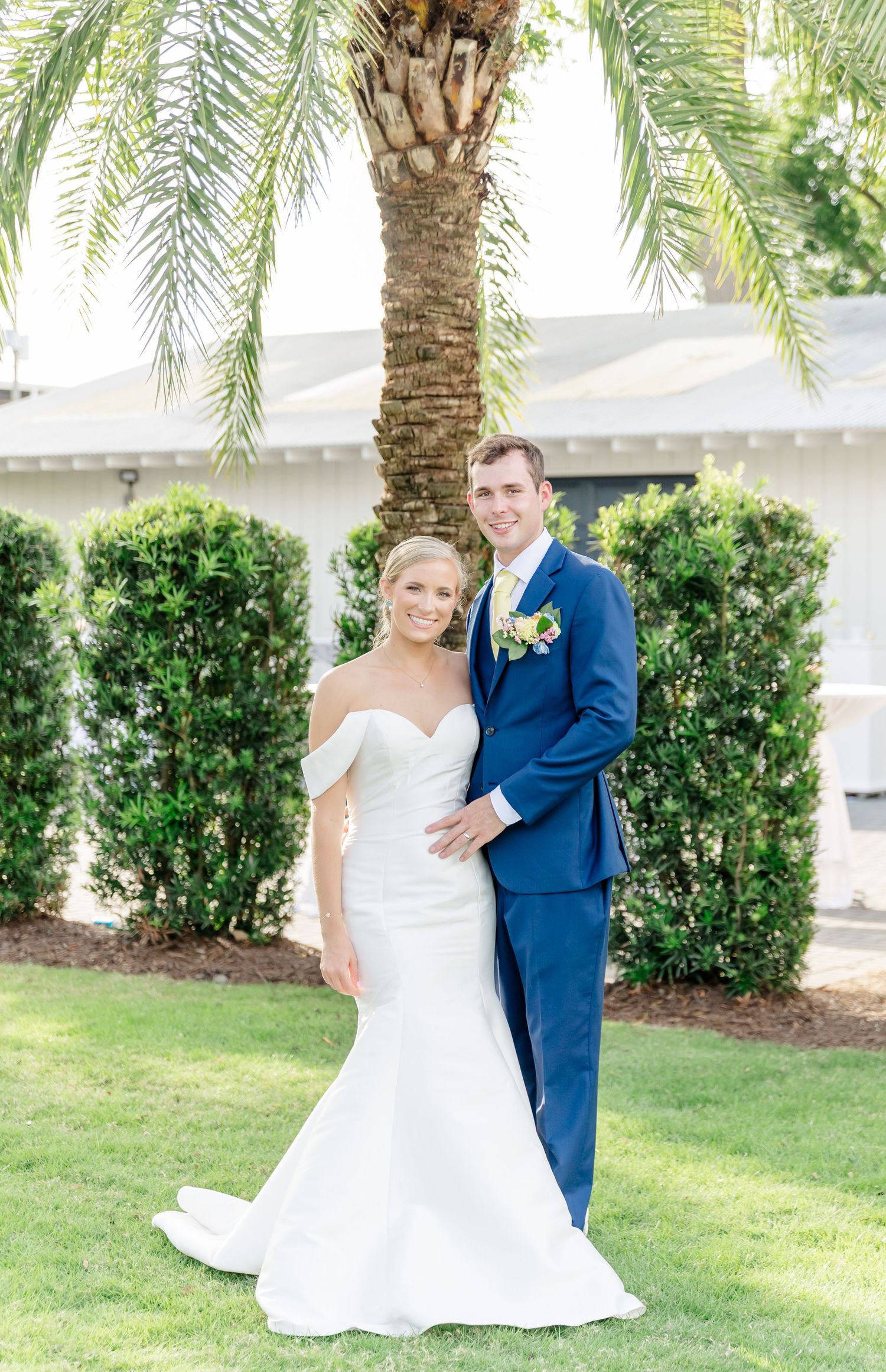 Newlyweds pose together outdoors on their wedding day surrounded by lush greenery and palm trees.