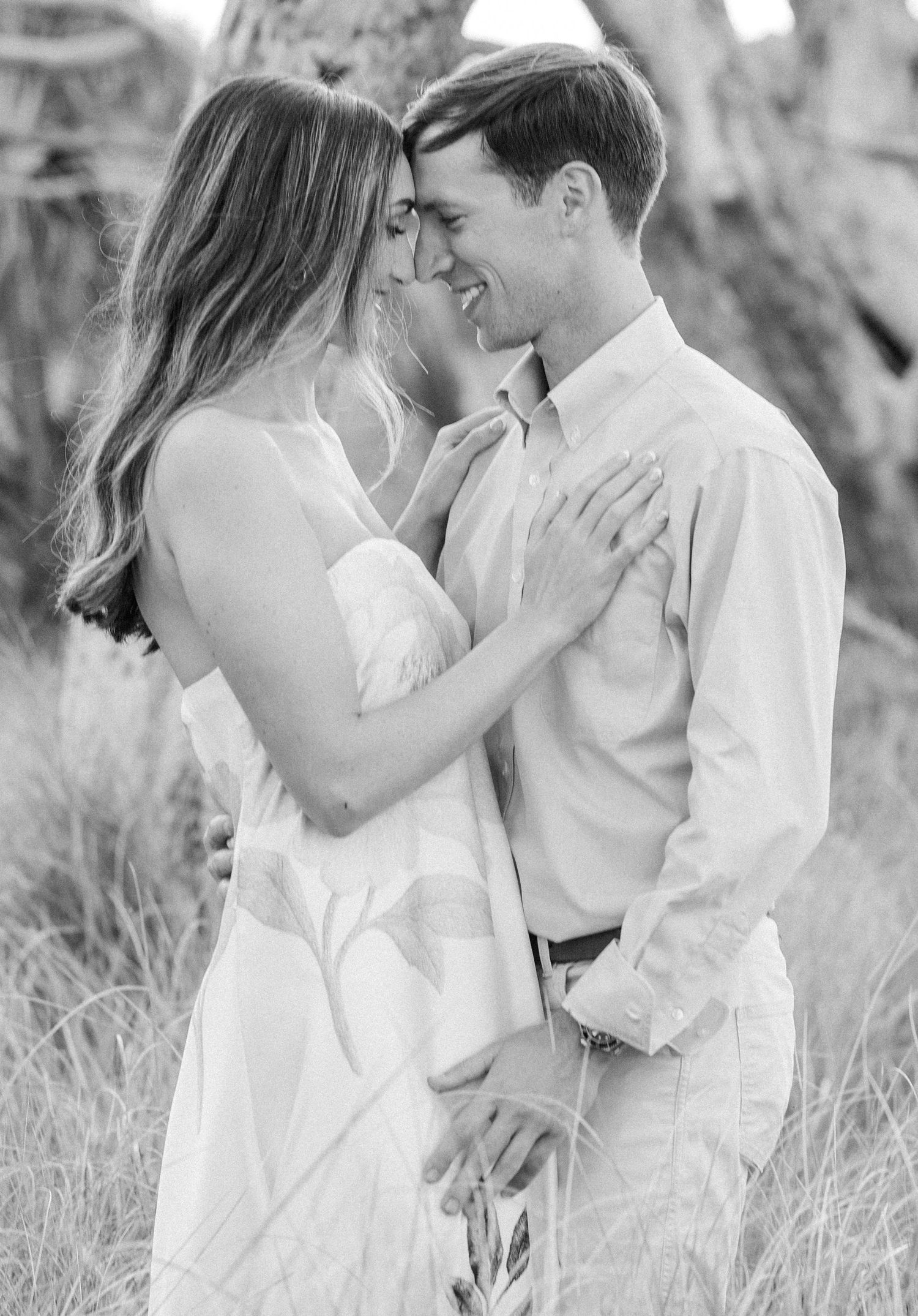 Black and white romantic photo of a smiling couple embracing closely in a field of tall grass.