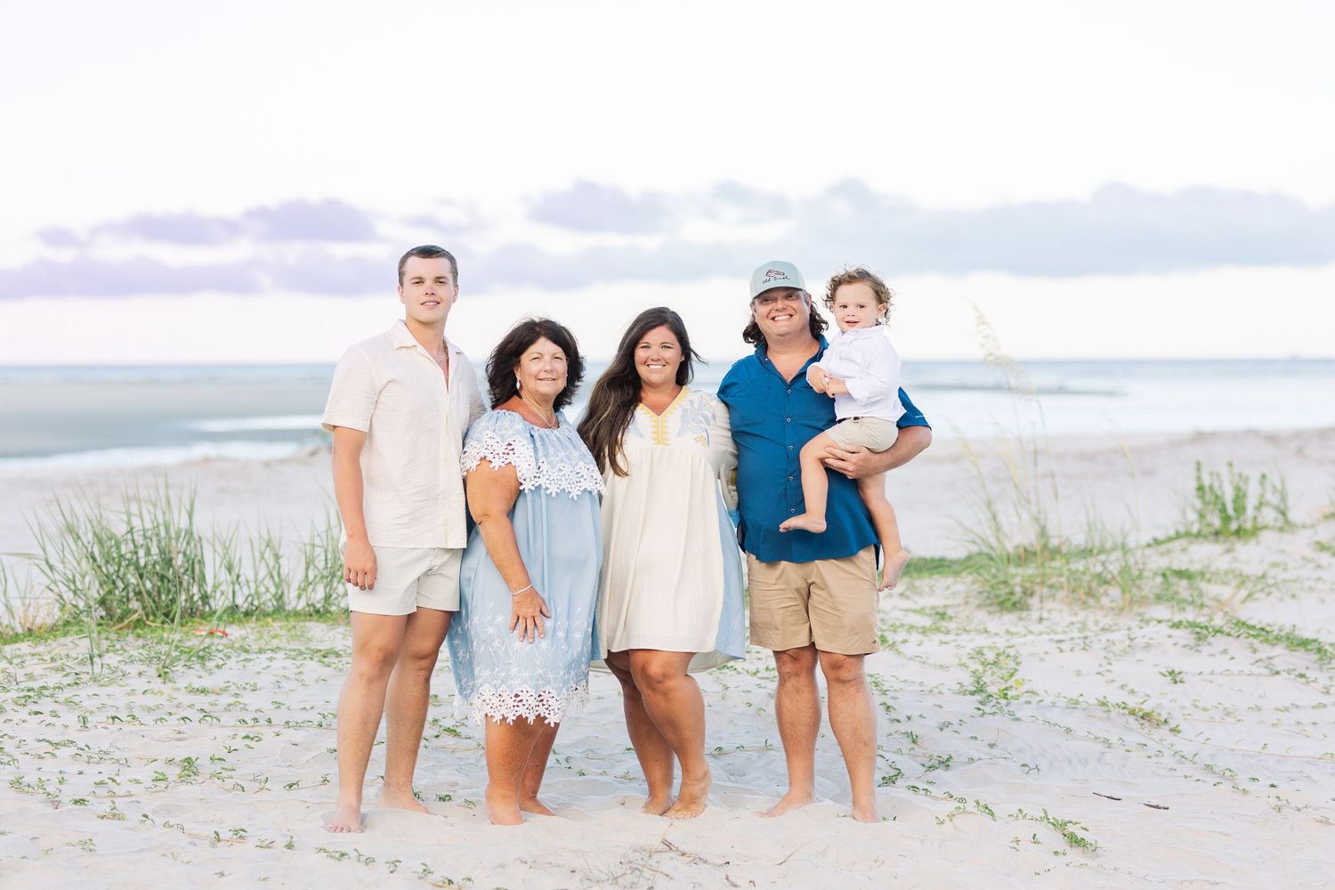 Five people in light-colored clothing stand together on a sandy beach at sunset with ocean waves in the background.