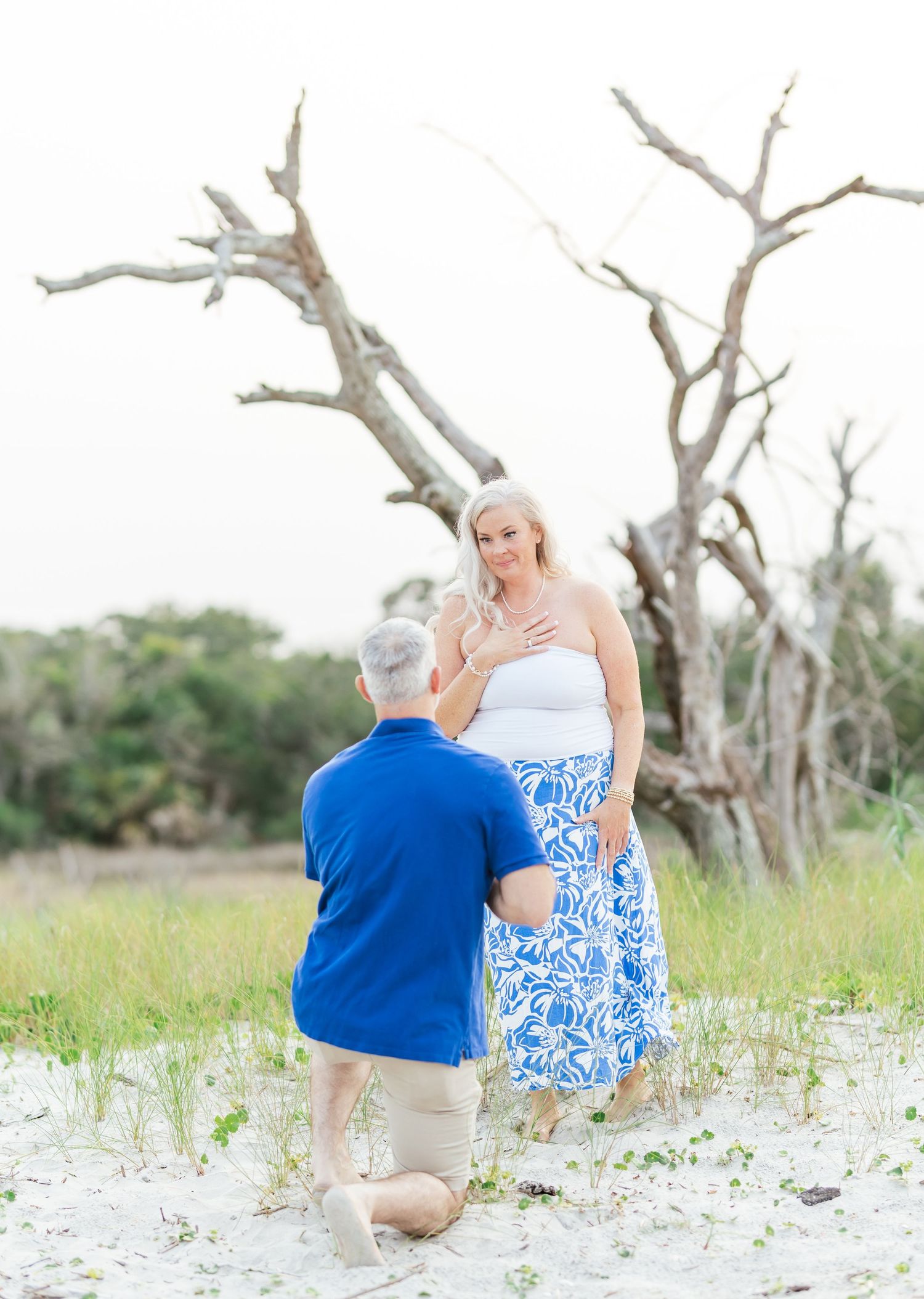 A proposal moment captured on a beach with bare trees in the background.