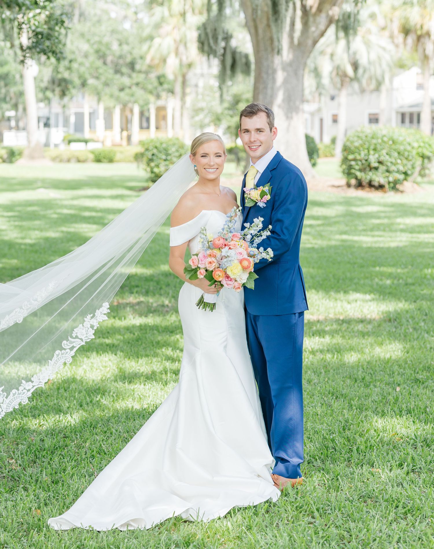 A wedding couple poses together outdoors in a garden setting with Spanish moss draped trees and lush greenery.