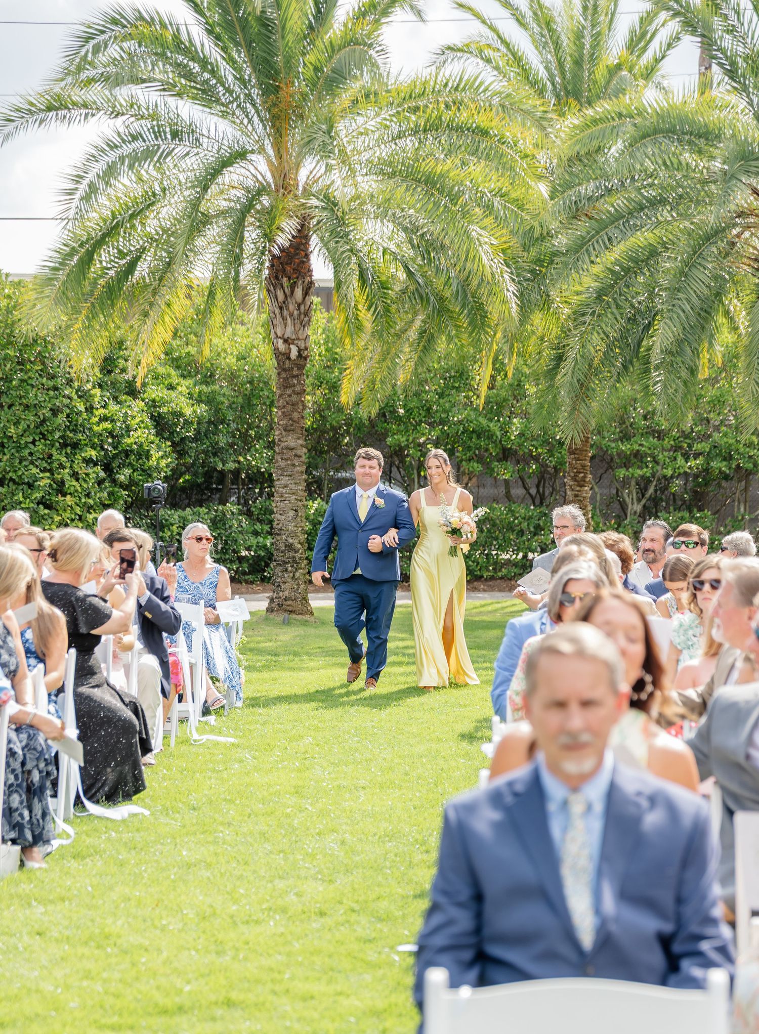 Wedding guests sit in rows of white chairs on a lush green lawn surrounded by towering palm trees during an outdoor ceremony.