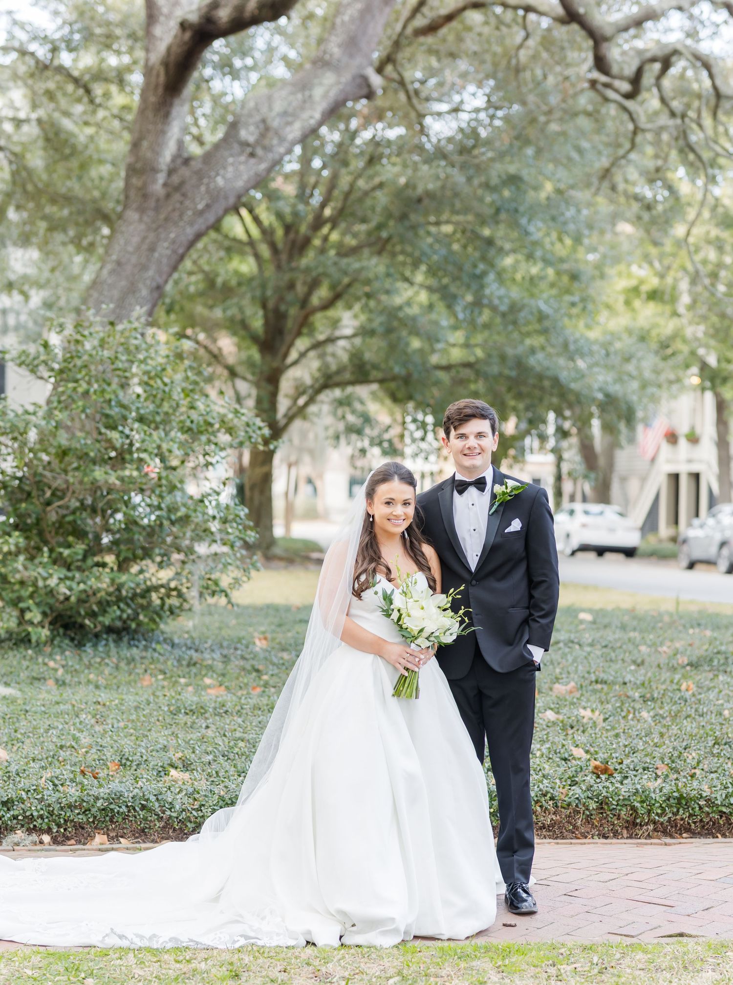 Elegant wedding couple poses outdoors under oak trees on a brick pathway.