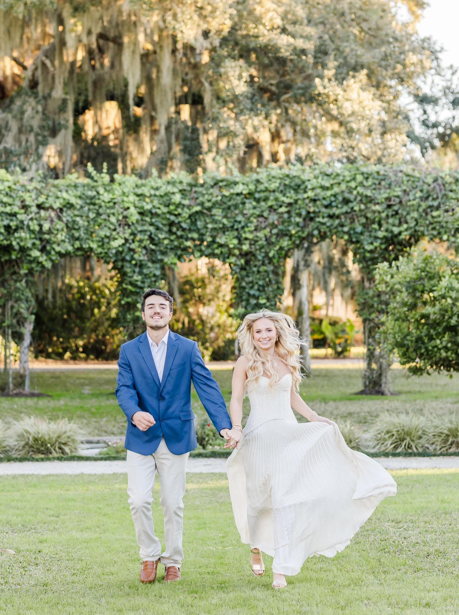 Couple playfully running and dancing together in a sun-dappled garden with Spanish moss draped trees.