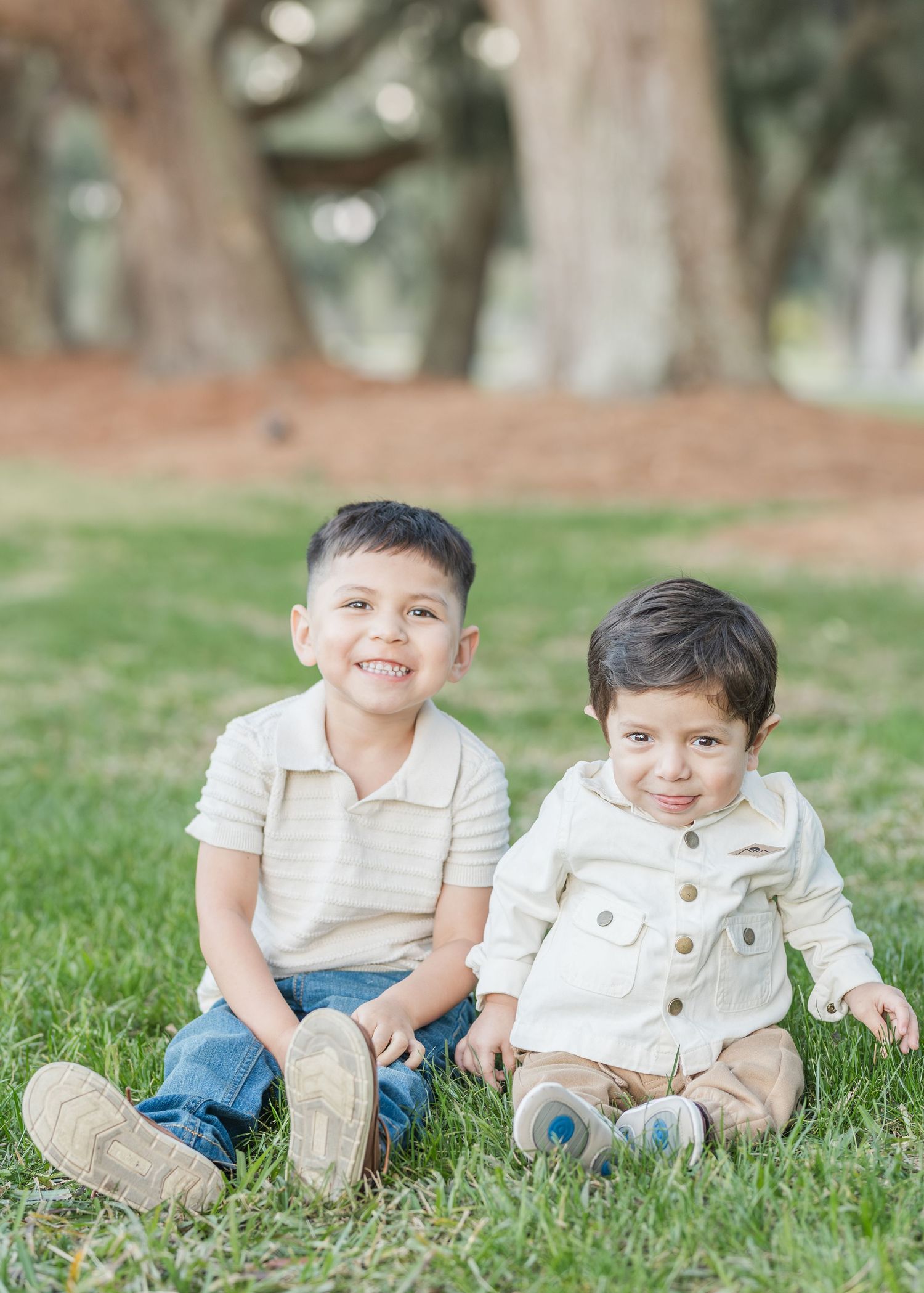 Two children sitting together on grass in coordinated white and neutral colored outfits in an outdoor setting.