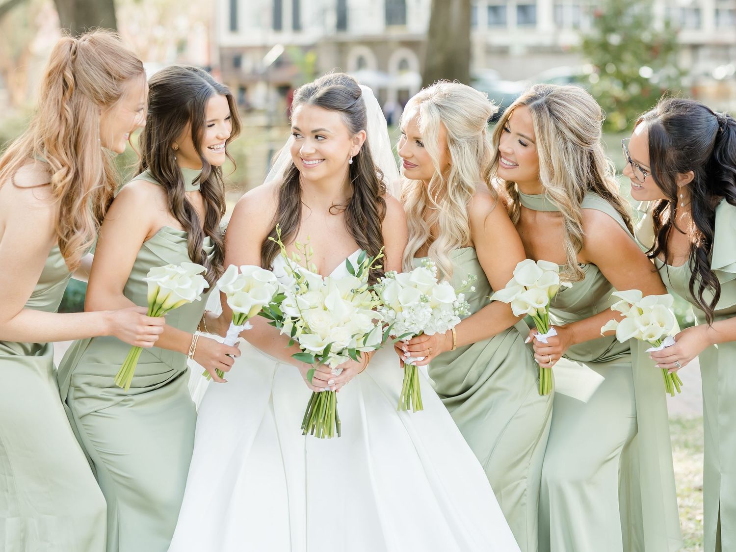 Bridesmaids in sage green dresses hold white bouquets while standing together outdoors.