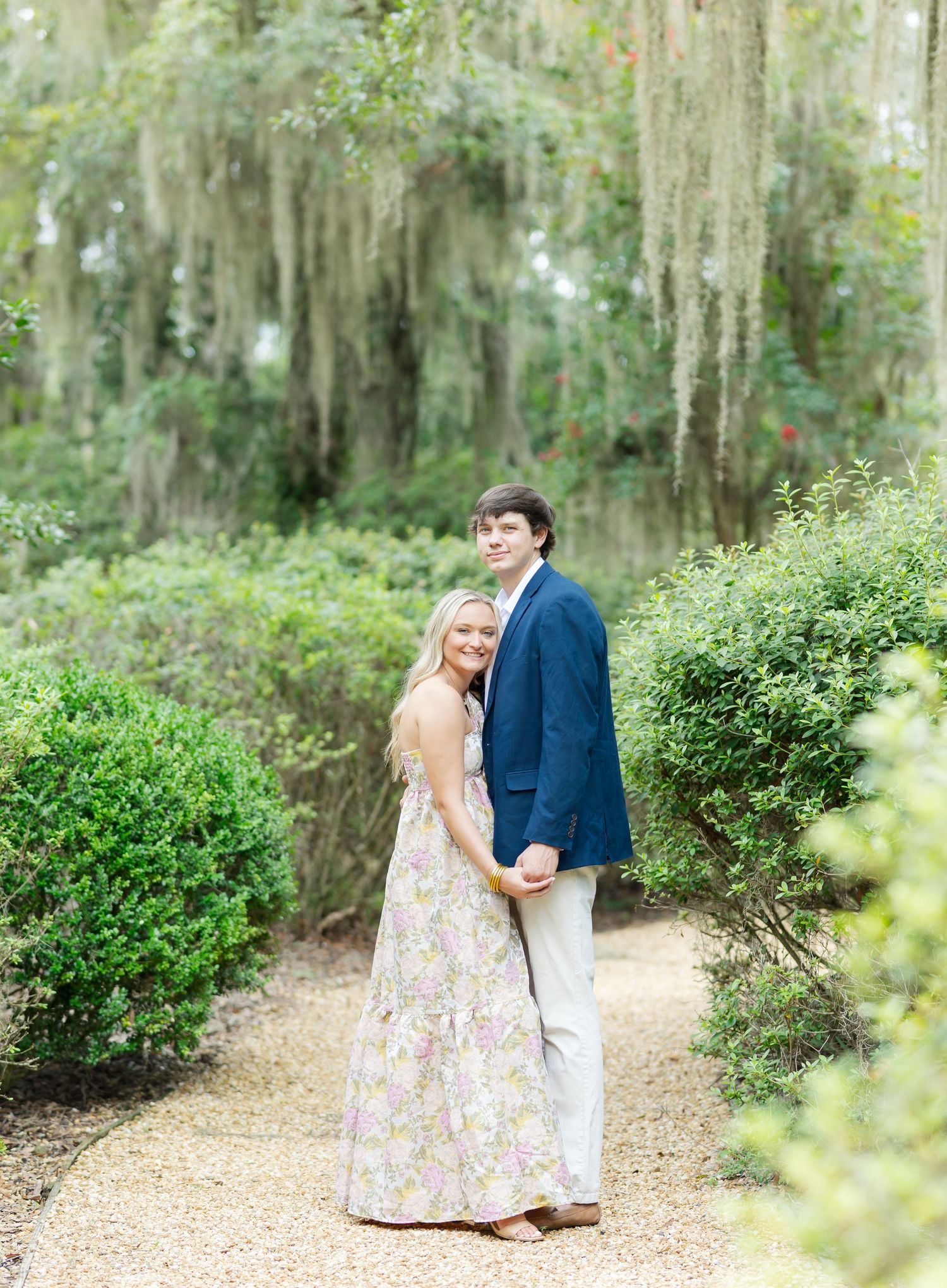 Romantic couple poses along a scenic garden pathway with hanging moss and green foliage.