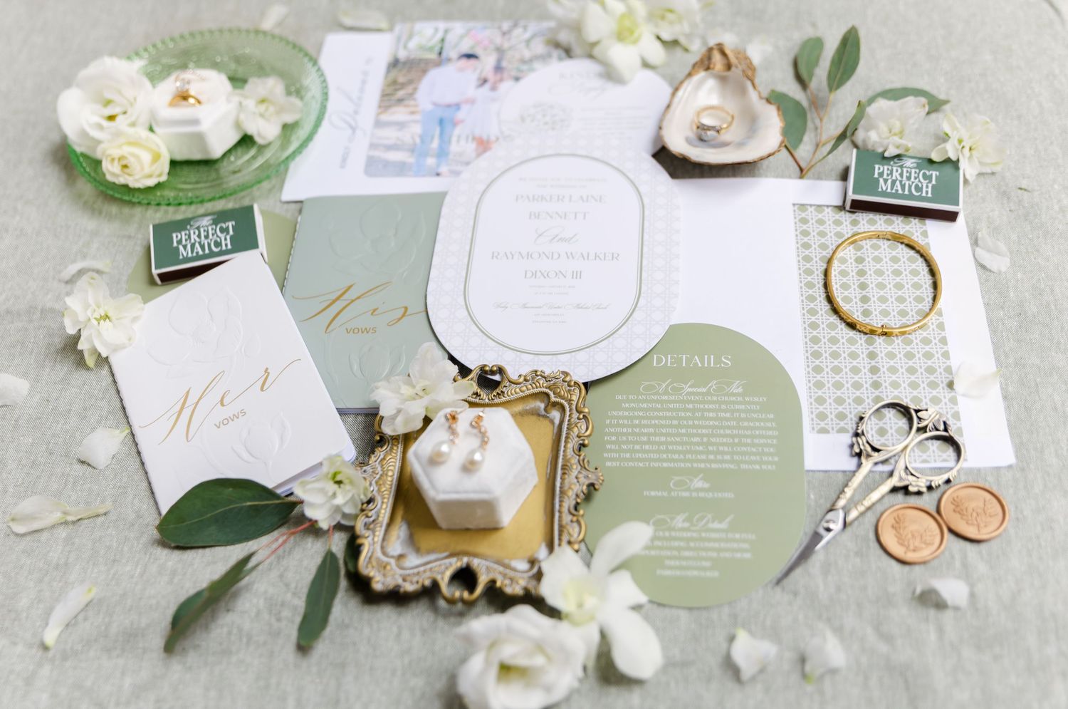 Wedding stationery flat lay with sage green invitations, rings, and white floral accents.
