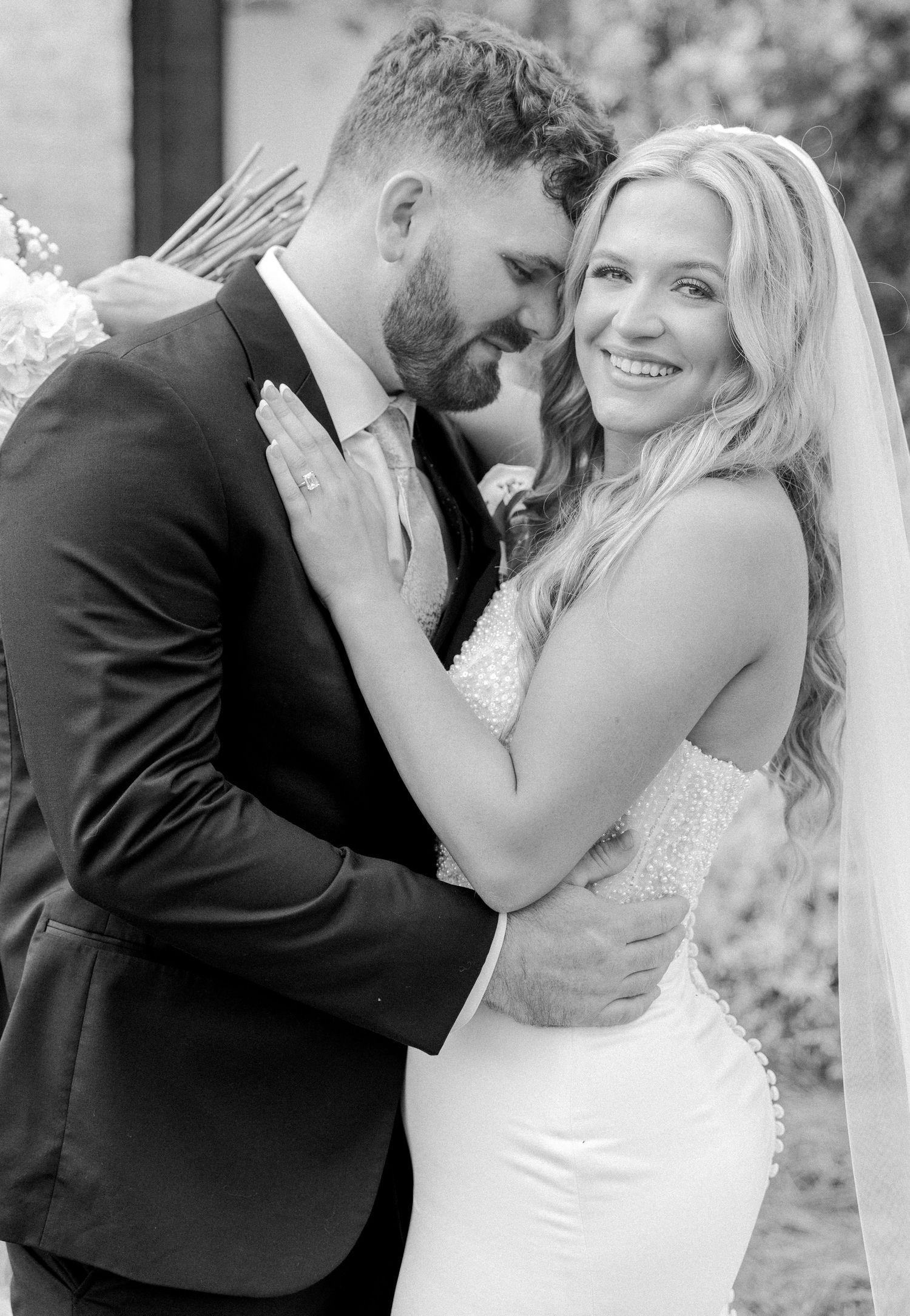 Black and white photo of smiling bride embracing groom in black suit at outdoor wedding venue.