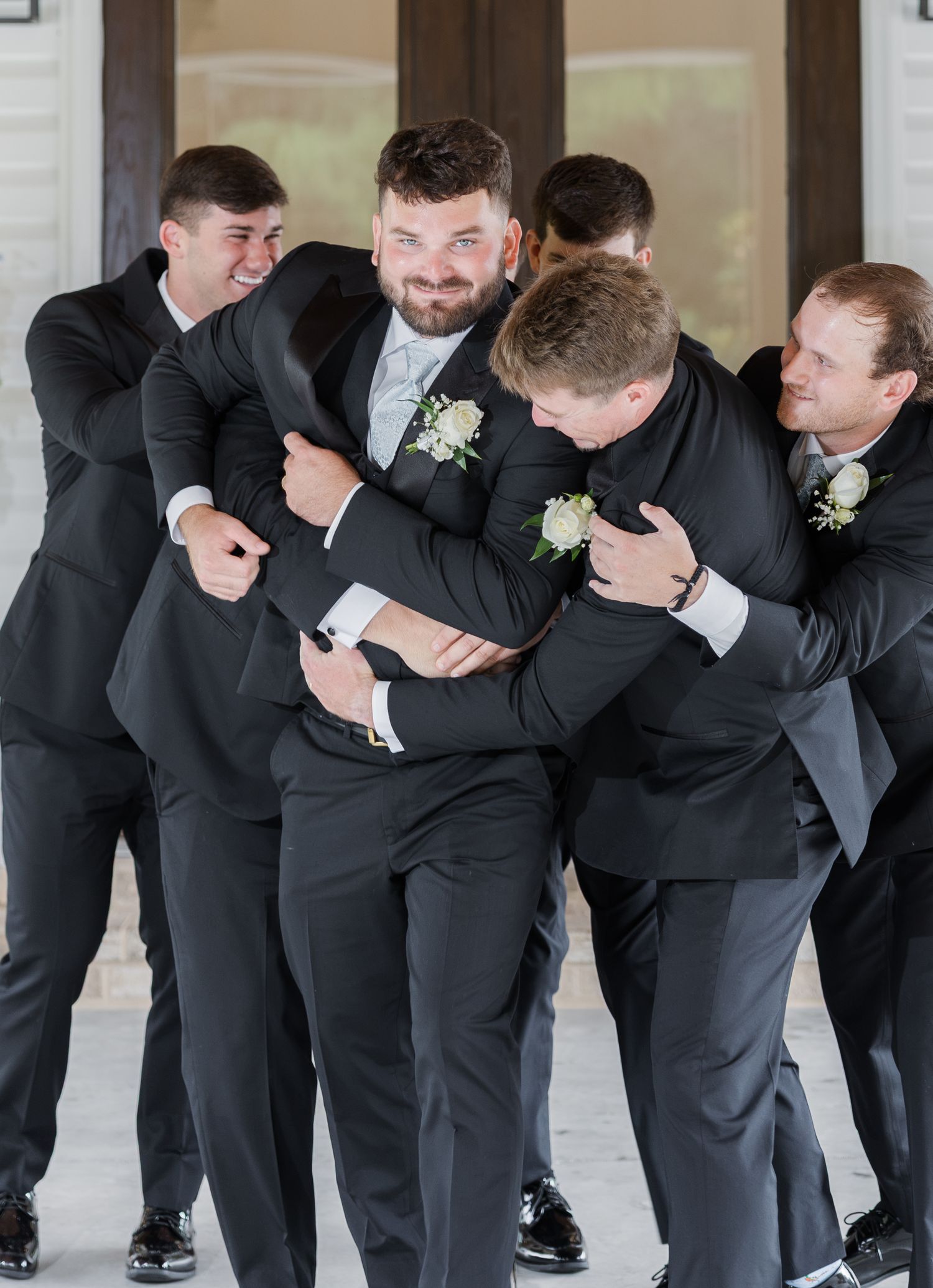 Groomsmen in black suits playfully piling onto the groom in a fun wedding party photo outdoors.