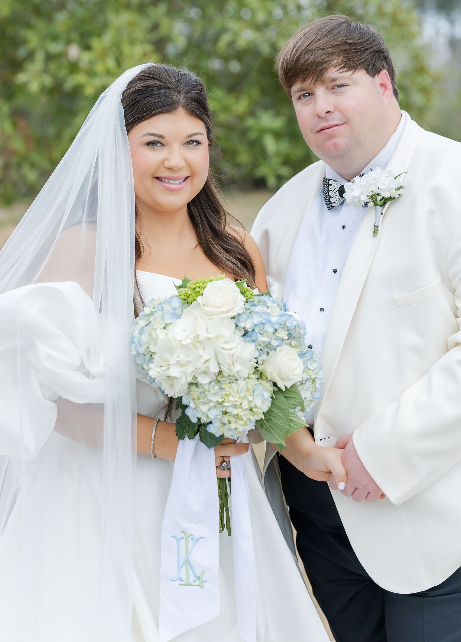 Smiling bride and groom posing outdoors with blue hydrangea and white rose bouquet with monogram ribbon on wedding day.
