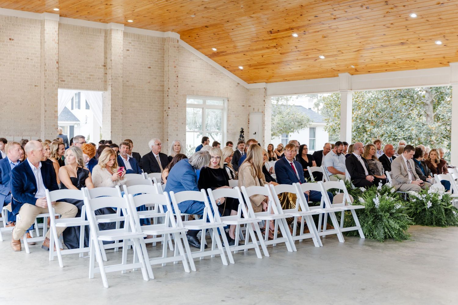 Wedding guests seated in white folding chairs inside bright open-air ceremony pavilion venue.