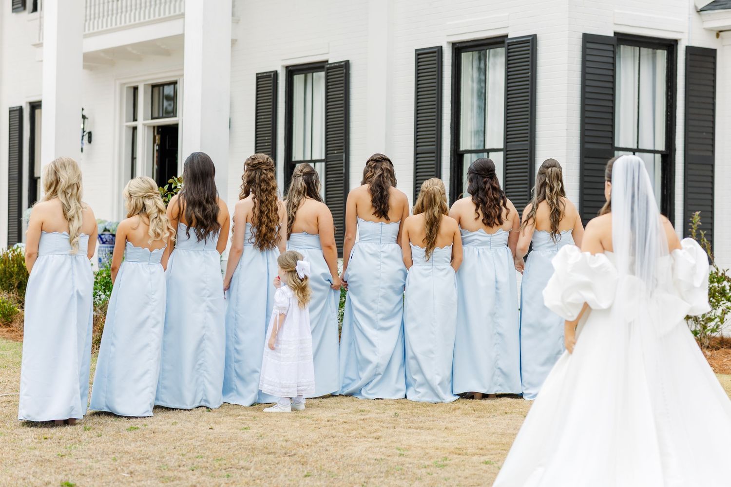 Bridesmaids in light blue gowns stand with backs to camera as bride approaches for first look reveal.