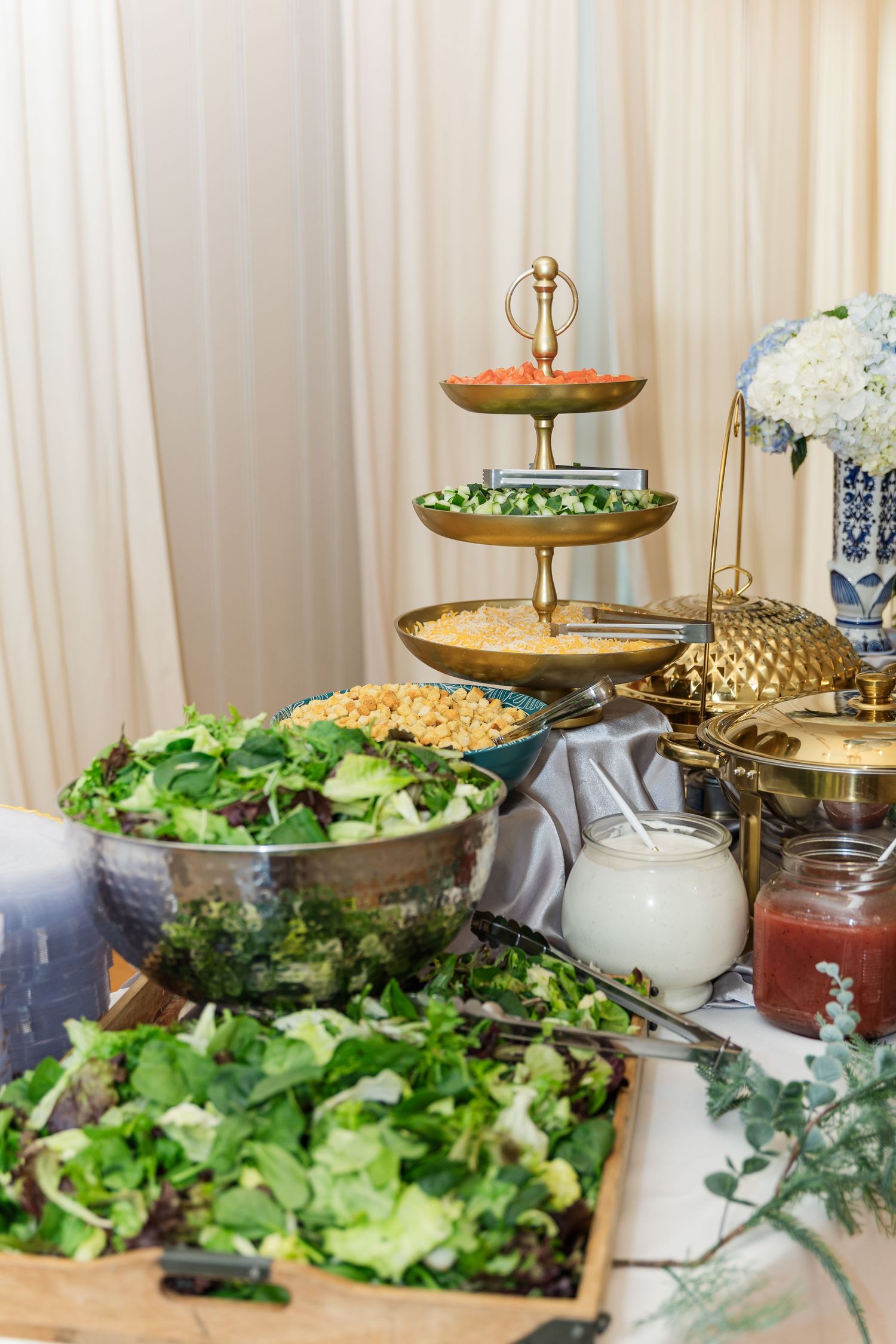Wedding reception buffet table with fresh green salads, gold chafing dishes, and tiered serving stand.