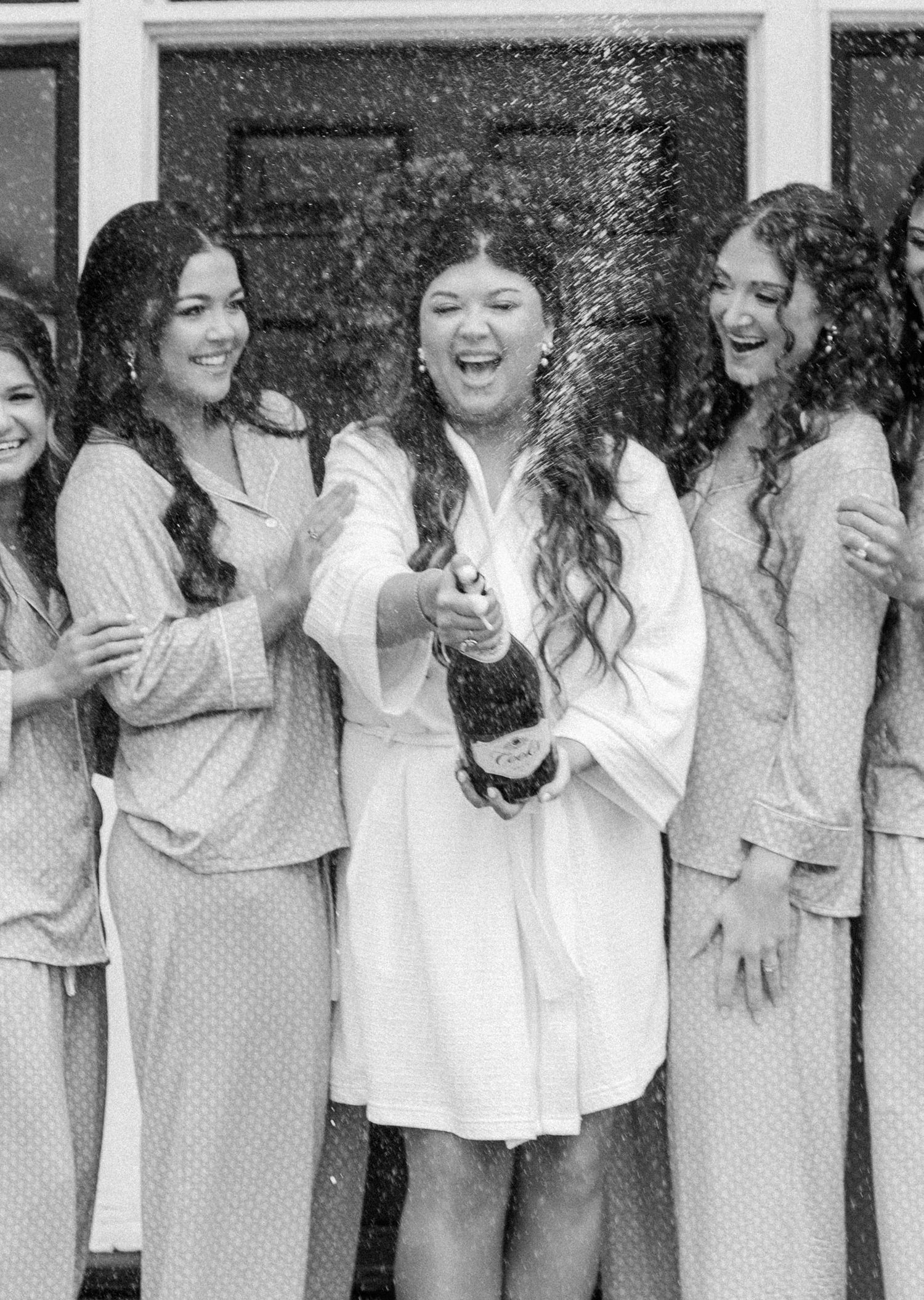 Black and white photo of bride popping champagne bottle surrounded by laughing bridesmaids in matching pink pajamas.