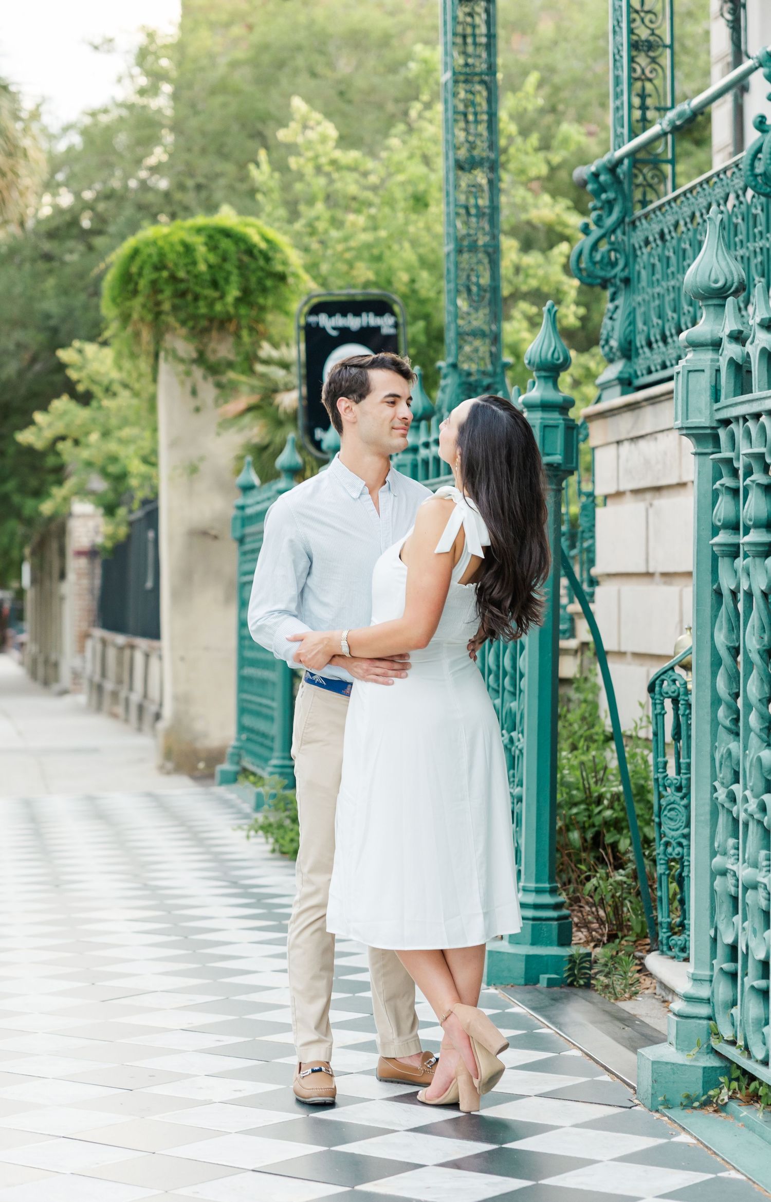 Couple in white dress and light blue shirt embracing by ornate teal iron gates on a checkered sidewalk.