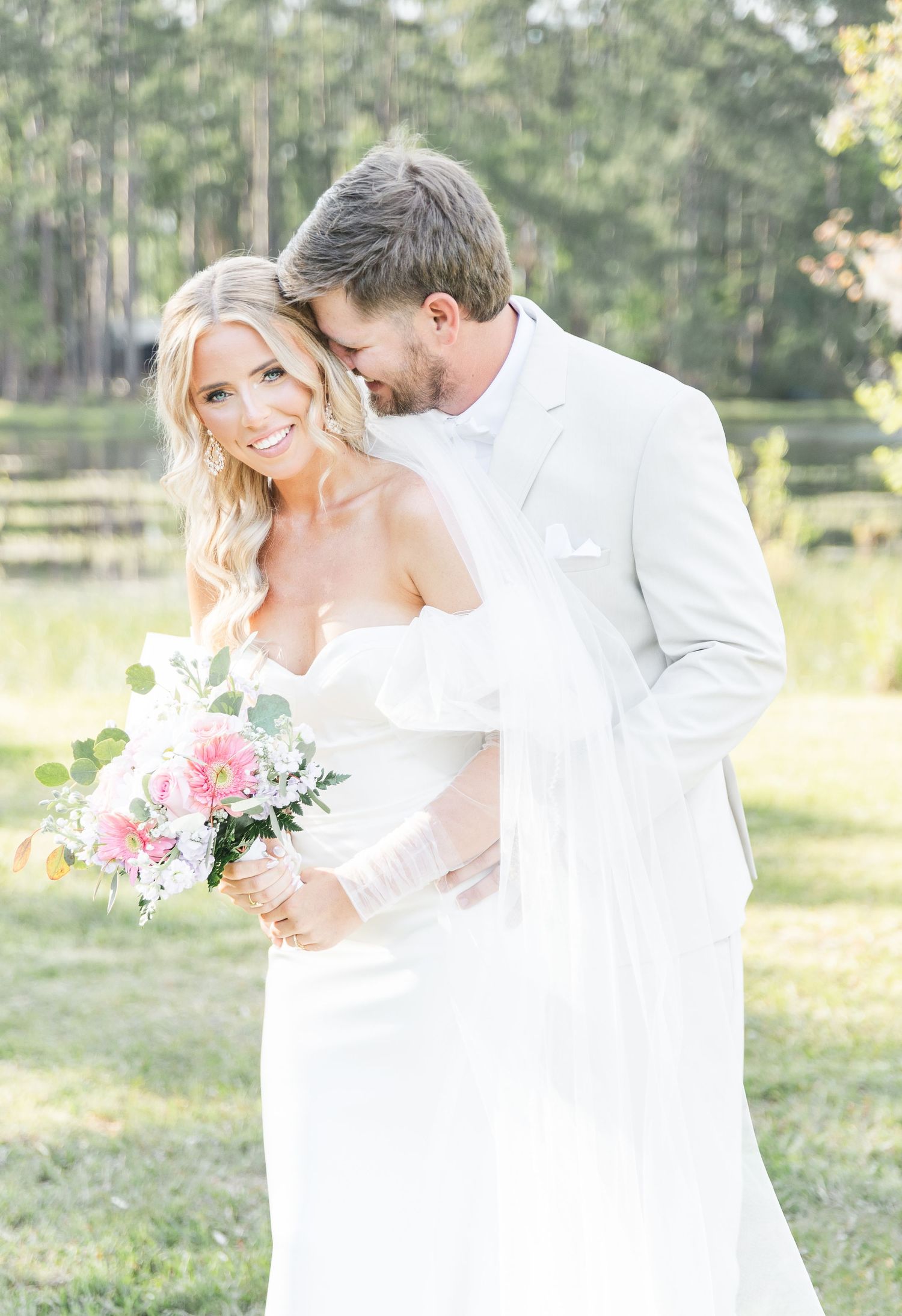 A joyful wedding couple embraces while holding pink floral bouquet in outdoor garden setting.