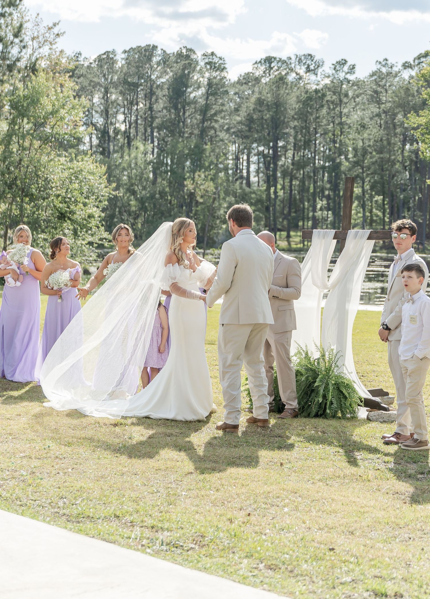 An outdoor wedding ceremony with bridesmaids in lavender dresses and groomsmen in light colored suits against a pine forest backdrop.