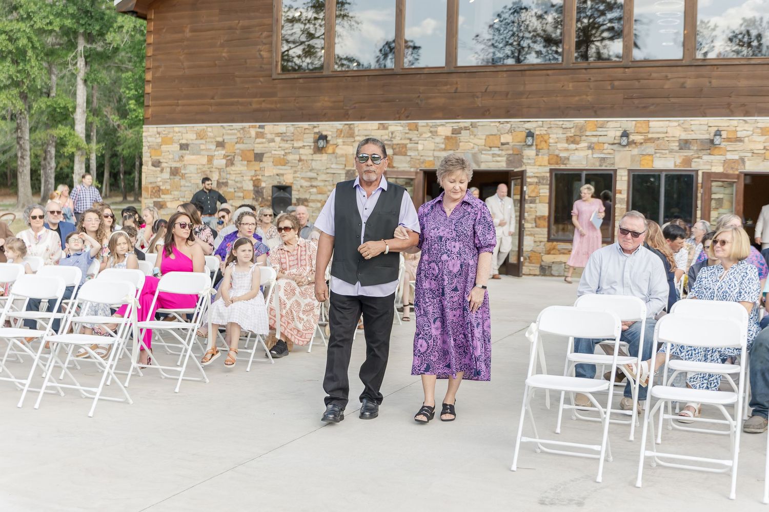 Wedding processional at rustic stone venue with guests seated in white folding chairs.