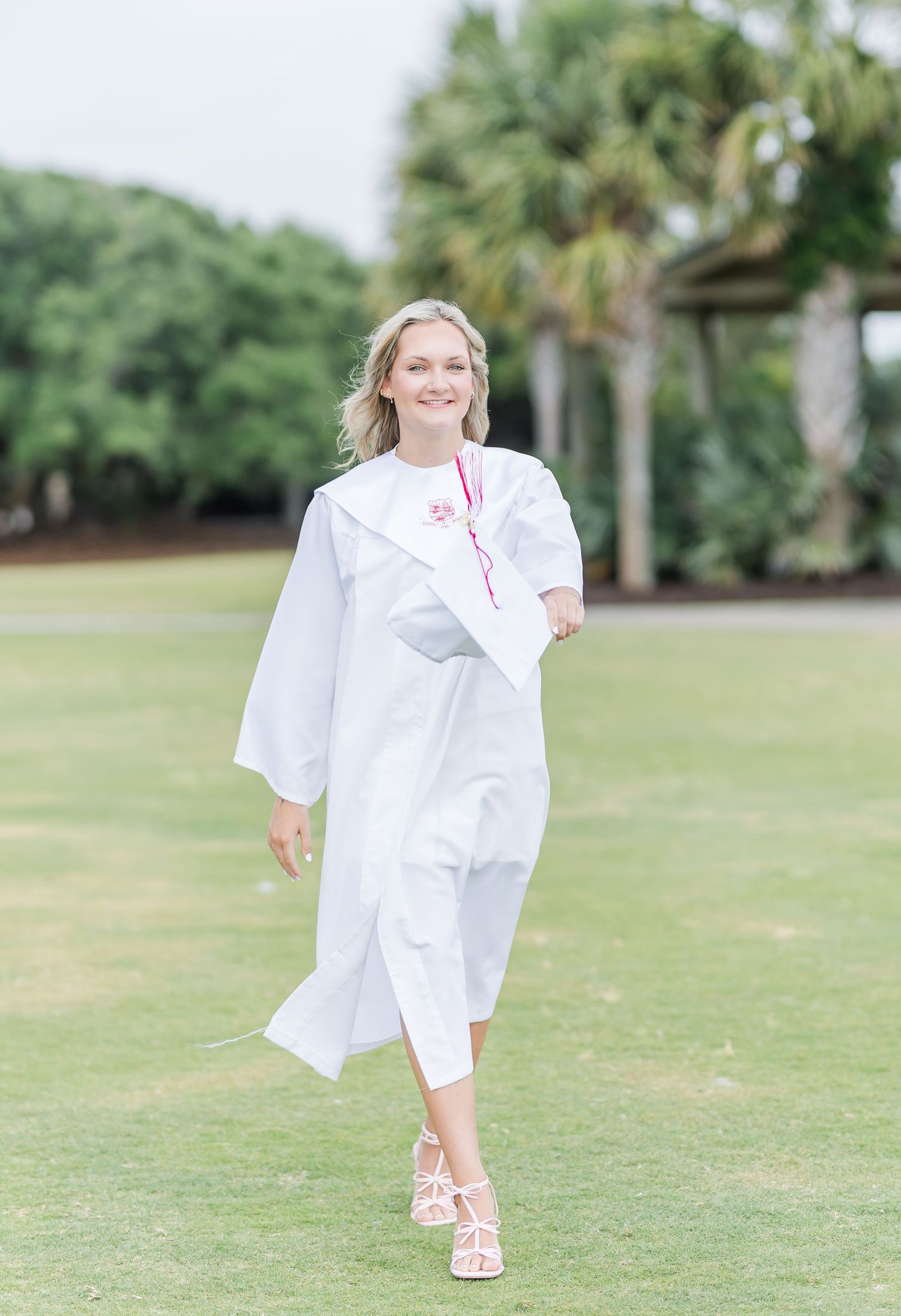 A person in white graduation attire walks confidently across a sunny lawn.