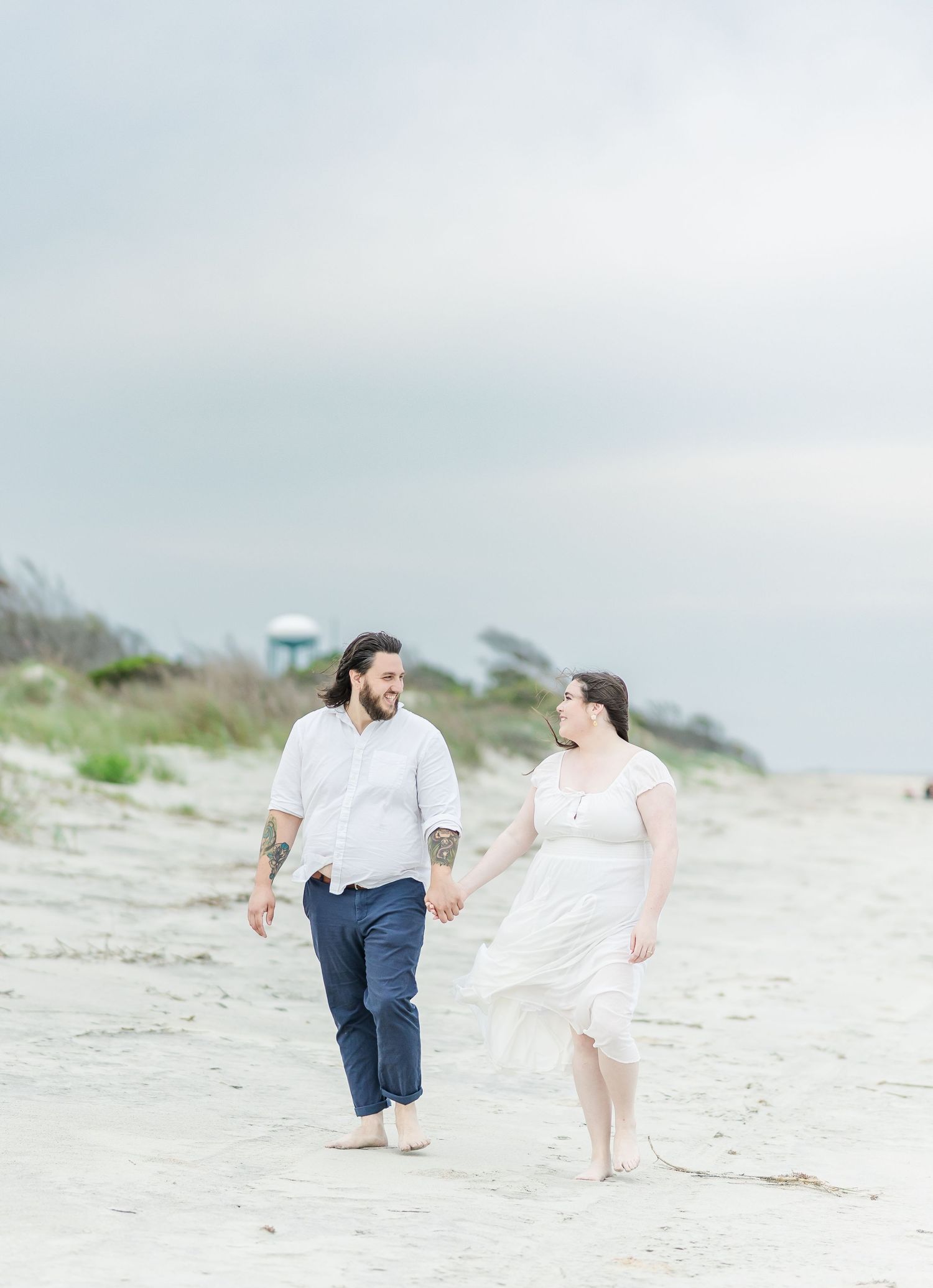 Couple walking barefoot on the beach holding hands in light airy clothing.