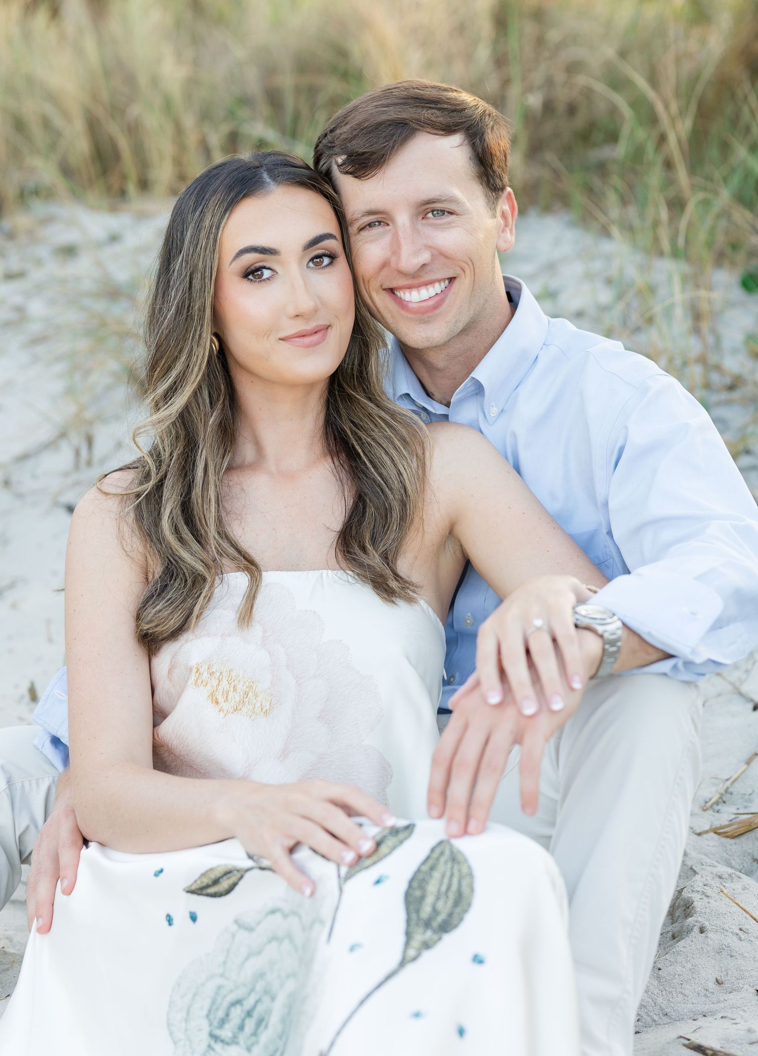 A smiling couple poses together on a sandy beach, the woman in a floral dress and the man in a light blue shirt.