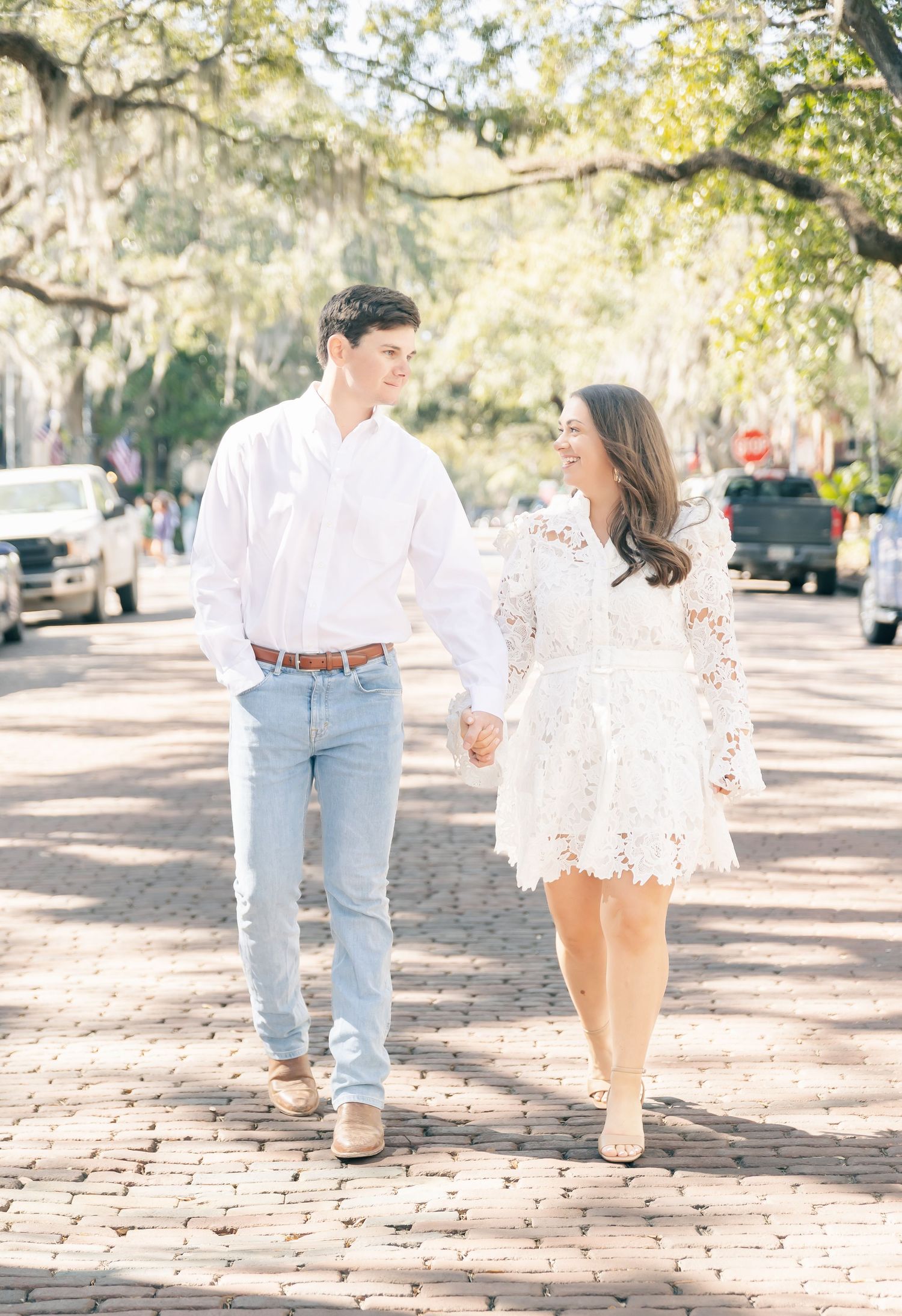 A couple strolls hand in hand down a charming brick-paved street with moss-draped trees.