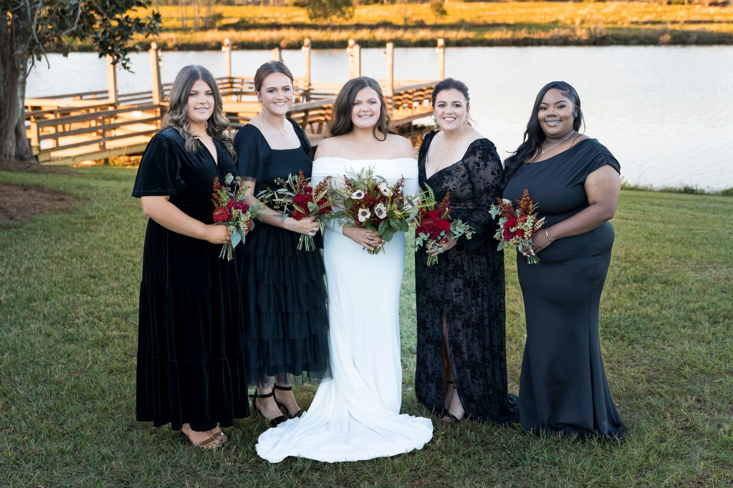 Wedding party in black formal attire poses together on a grassy lawn by the water.