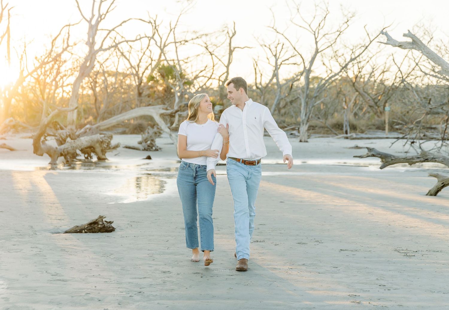 Casual couple walking together on beach with sunset and driftwood in background.