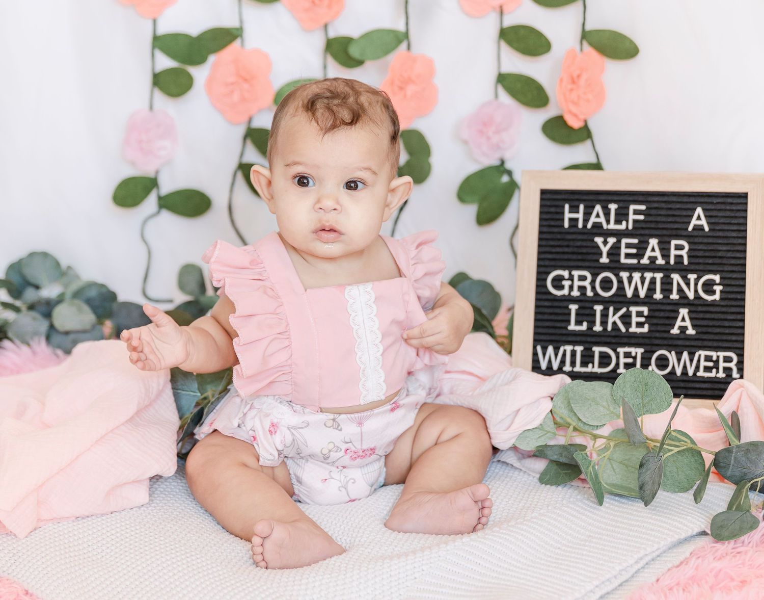 Baby in pink romper sits beside milestone sign with eucalyptus leaves and floral wall decor.