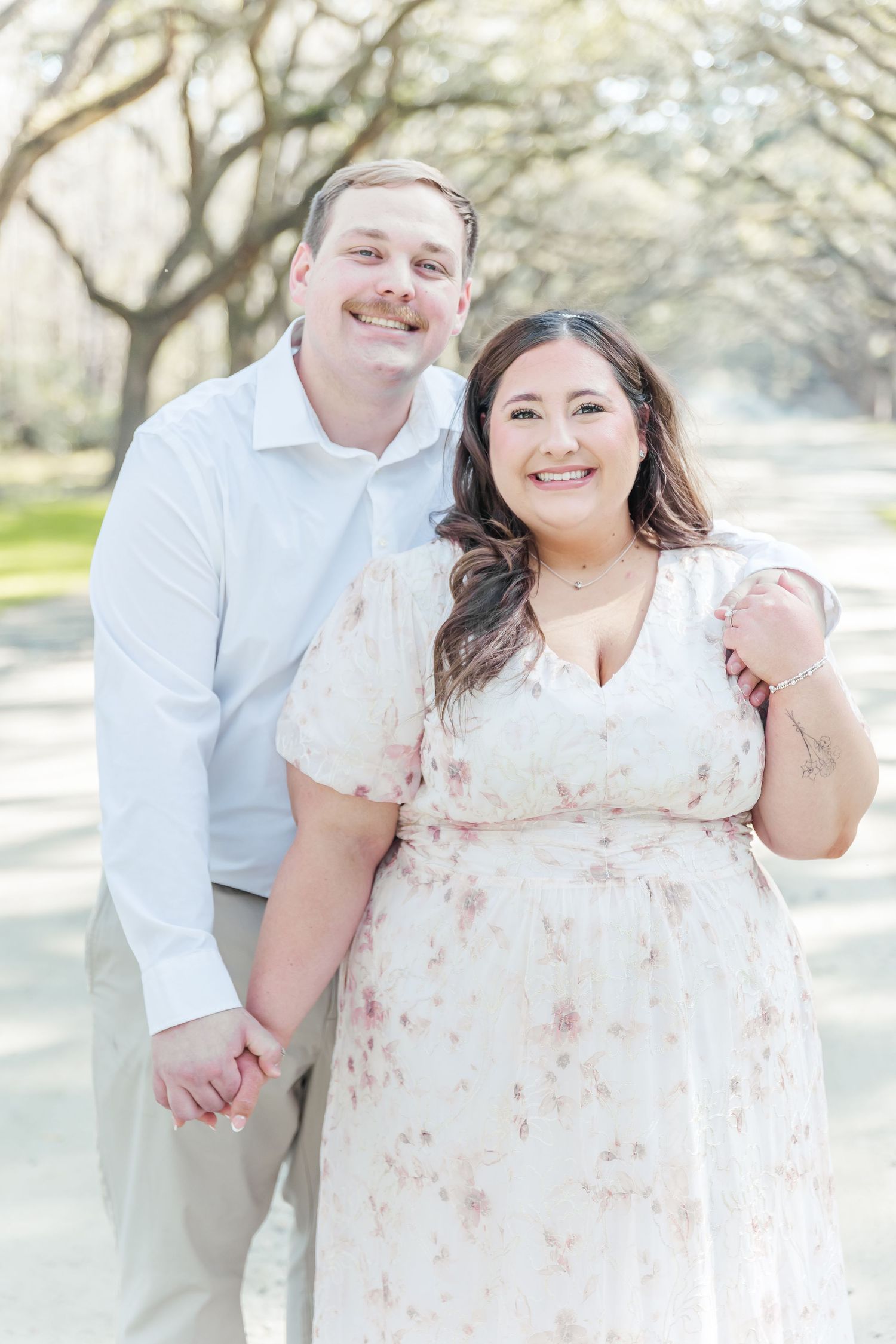 A romantic engagement photo under a canopy of oak trees features a couple in soft white and khaki attire.