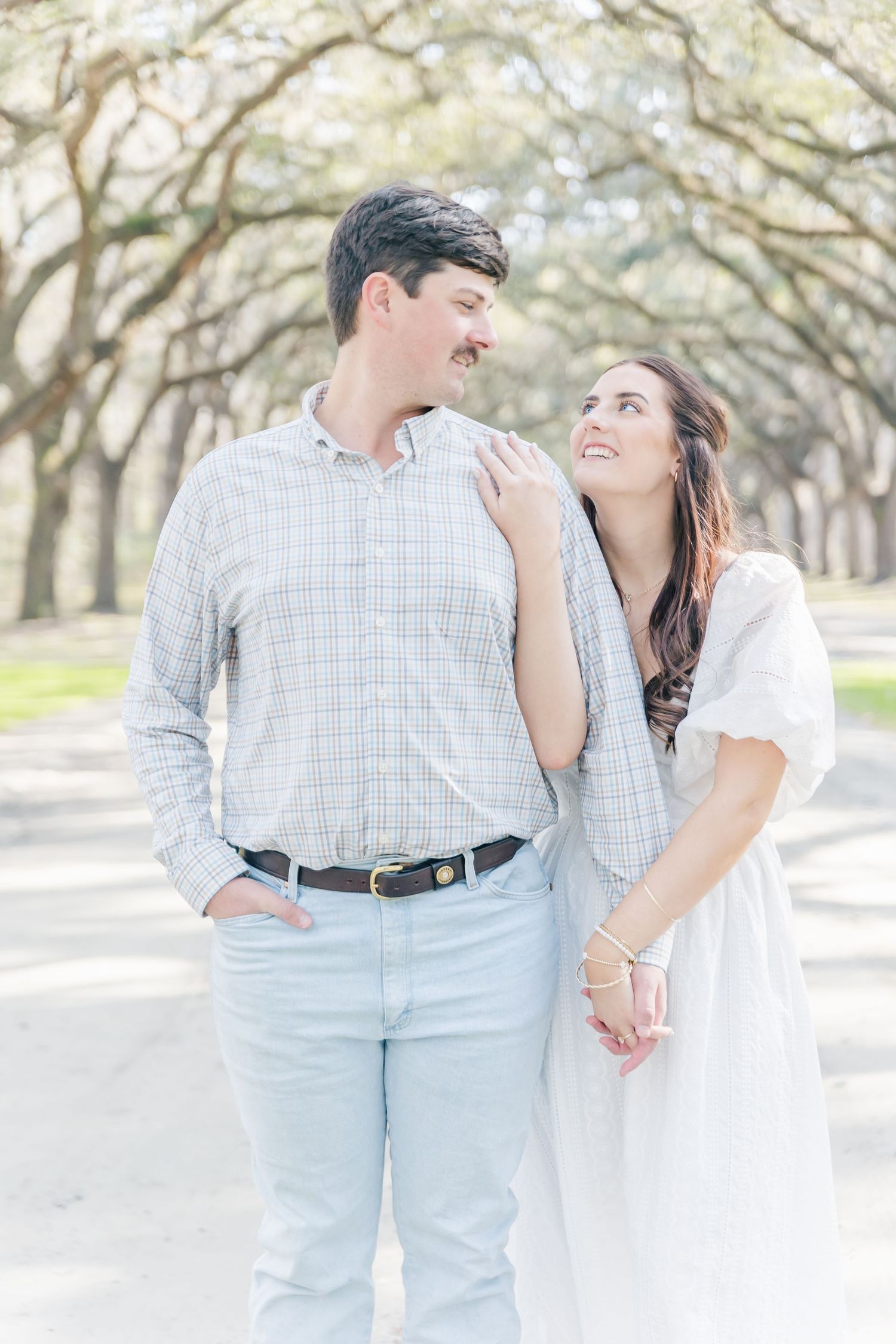 A couple embraces while standing on a path beneath arching oak trees.