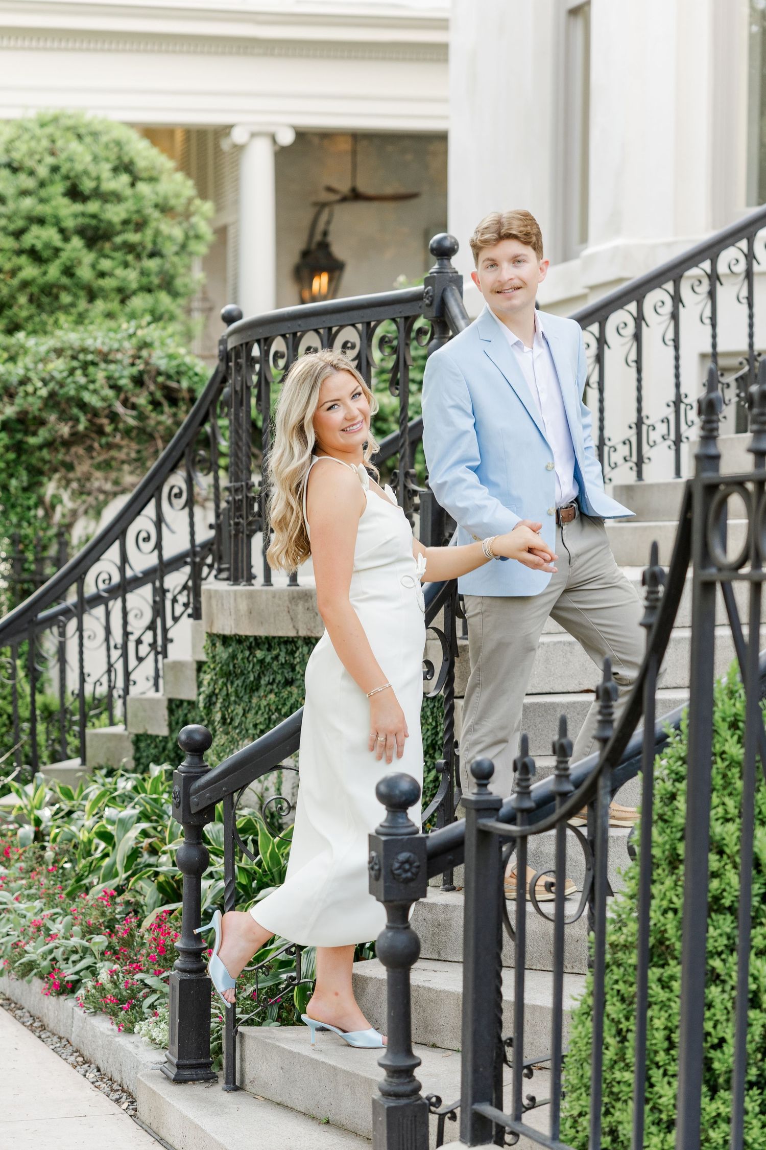 Couple posing on elegant iron staircase, woman in white dress and man in light blue blazer, smiling outdoors.