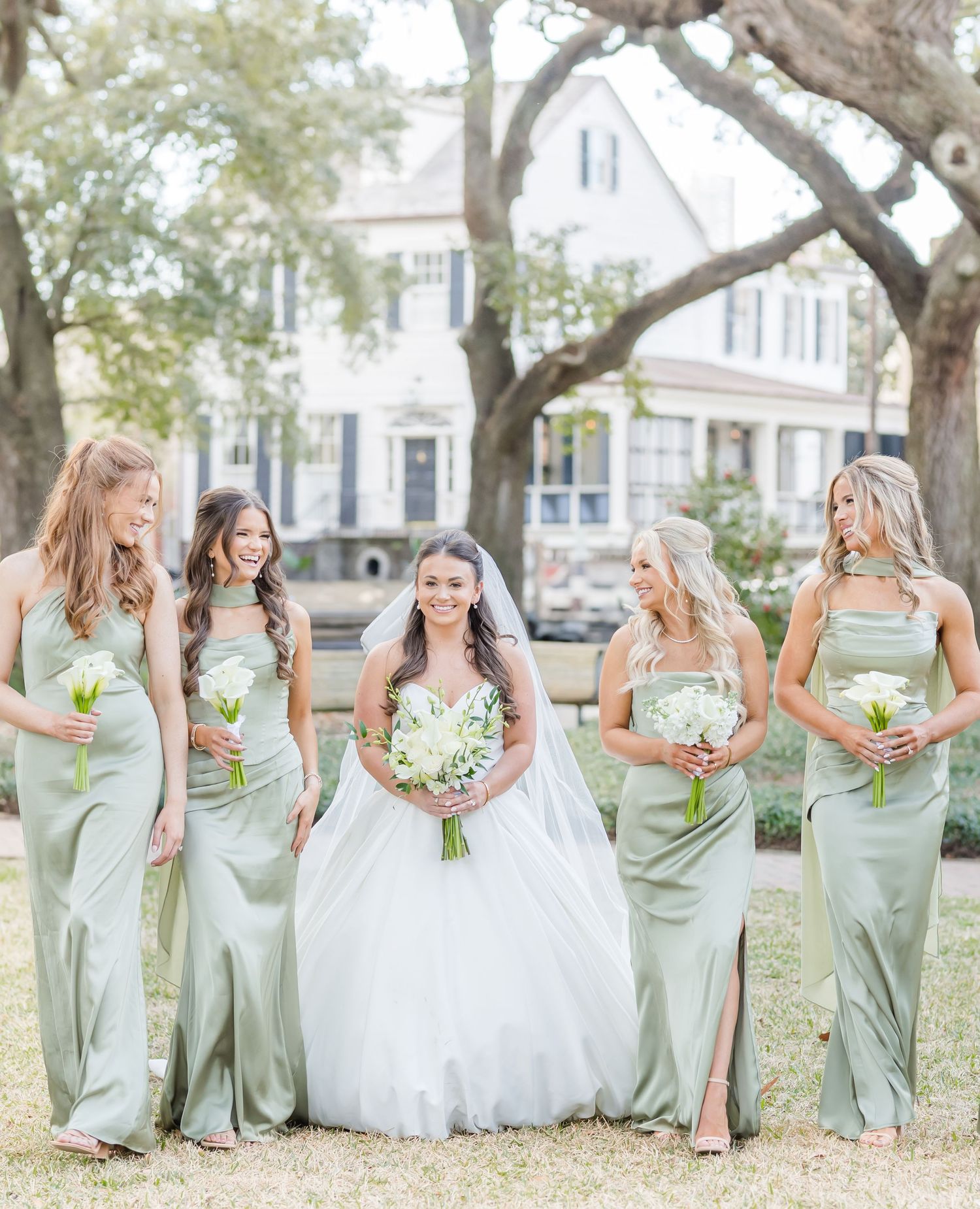 Bride surrounded by four bridesmaids in sage green gowns holding white bouquets outdoors.