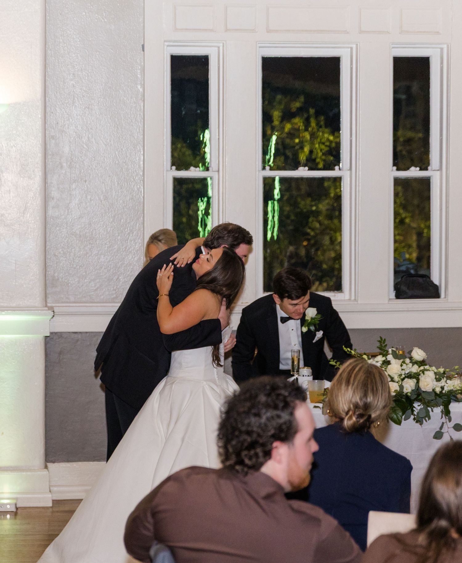 Bride and guest share a heartfelt hug at elegant wedding reception with floral centerpiece and guests seated nearby.