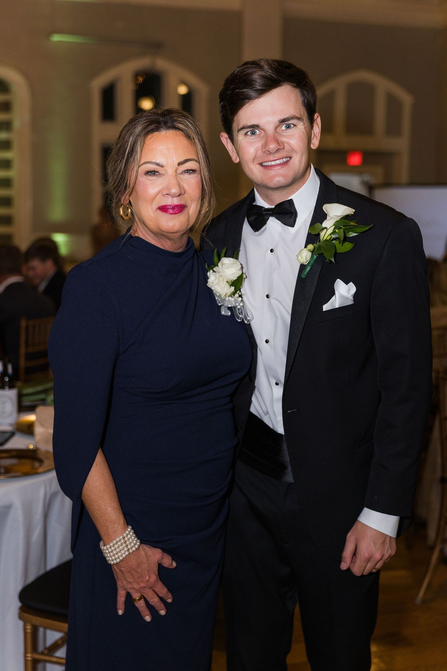 Woman in navy dress and young man in tuxedo posing together at wedding reception.