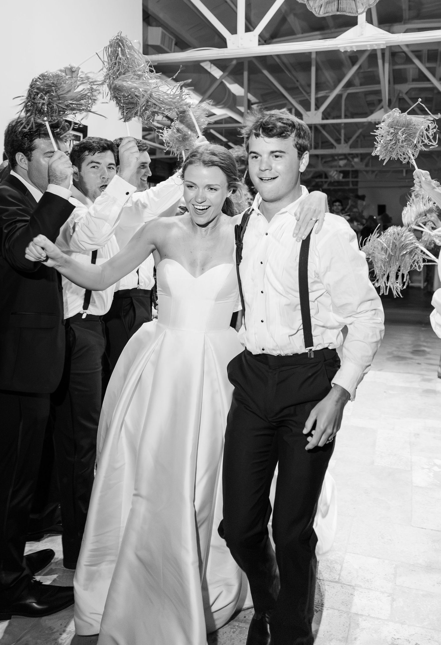 Smiling bride and groom exit reception in black and white photo with cheering guests.