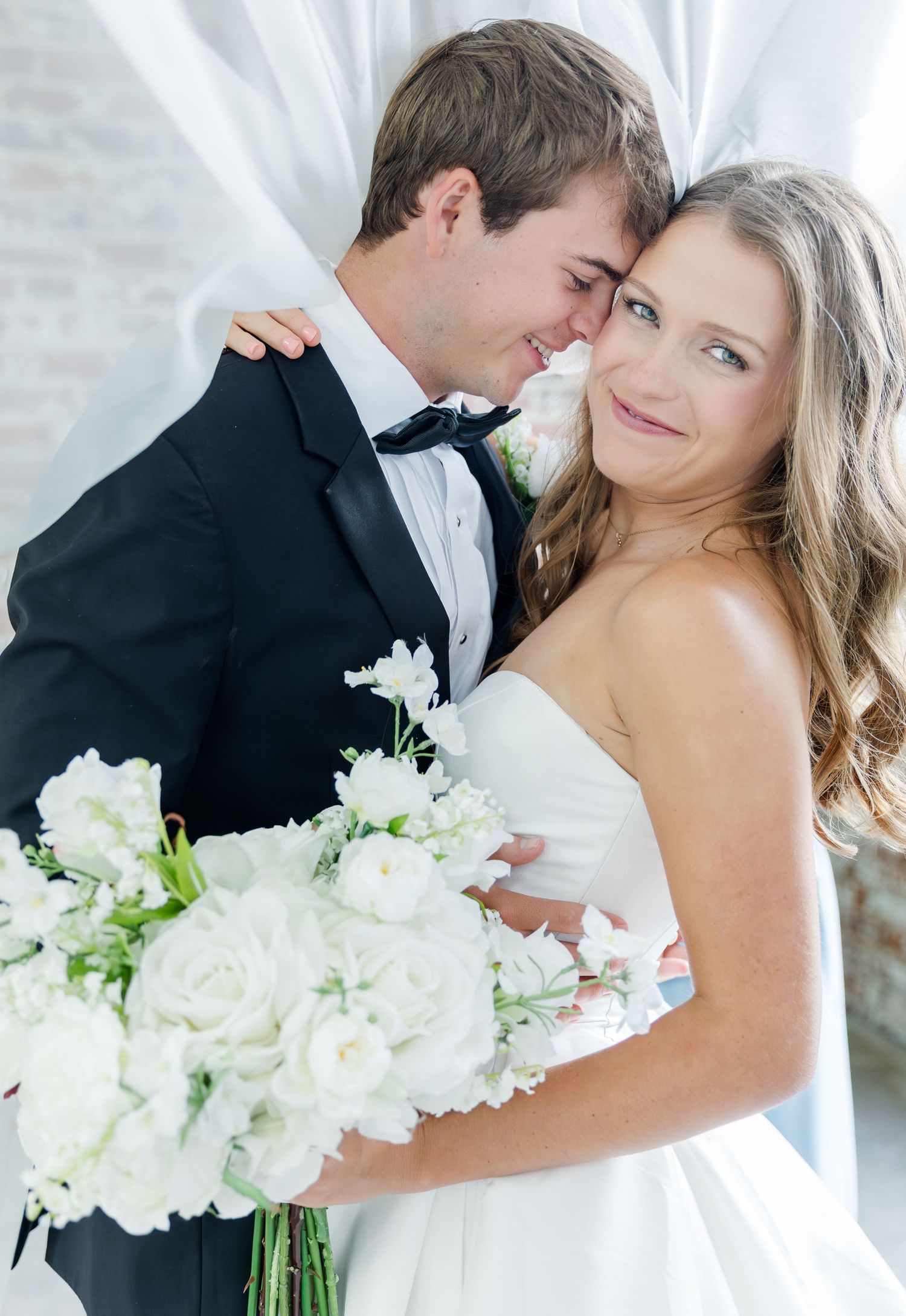 Bride holding white floral bouquet leans against groom in black tuxedo near sheer white curtains.