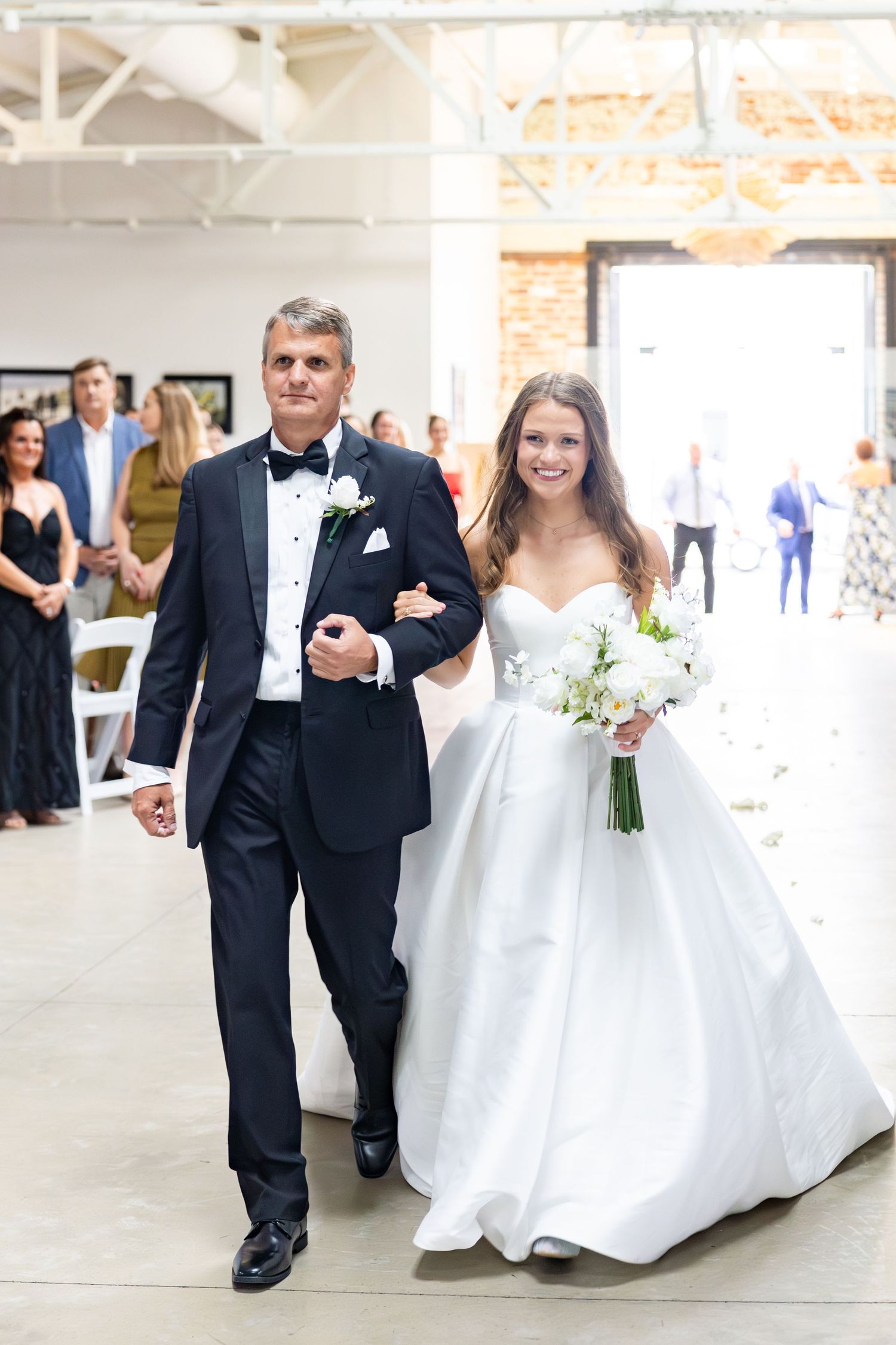 Father escorts radiant bride in white ball gown down aisle at bright industrial wedding venue.
