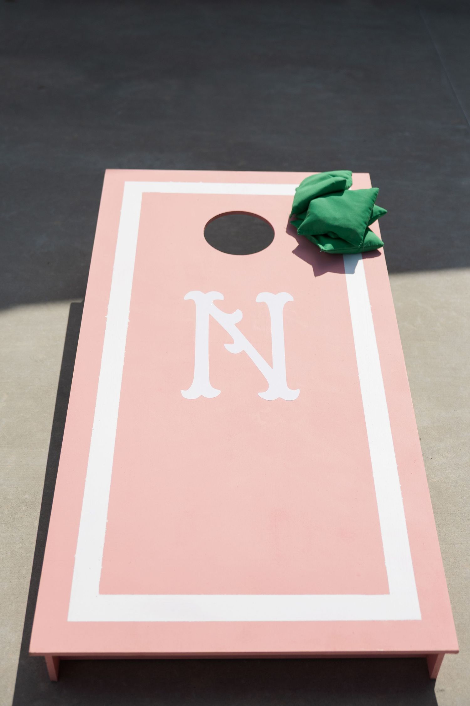 Pink monogrammed cornhole board with a green bean bag on a gray pavement outdoor wedding game.