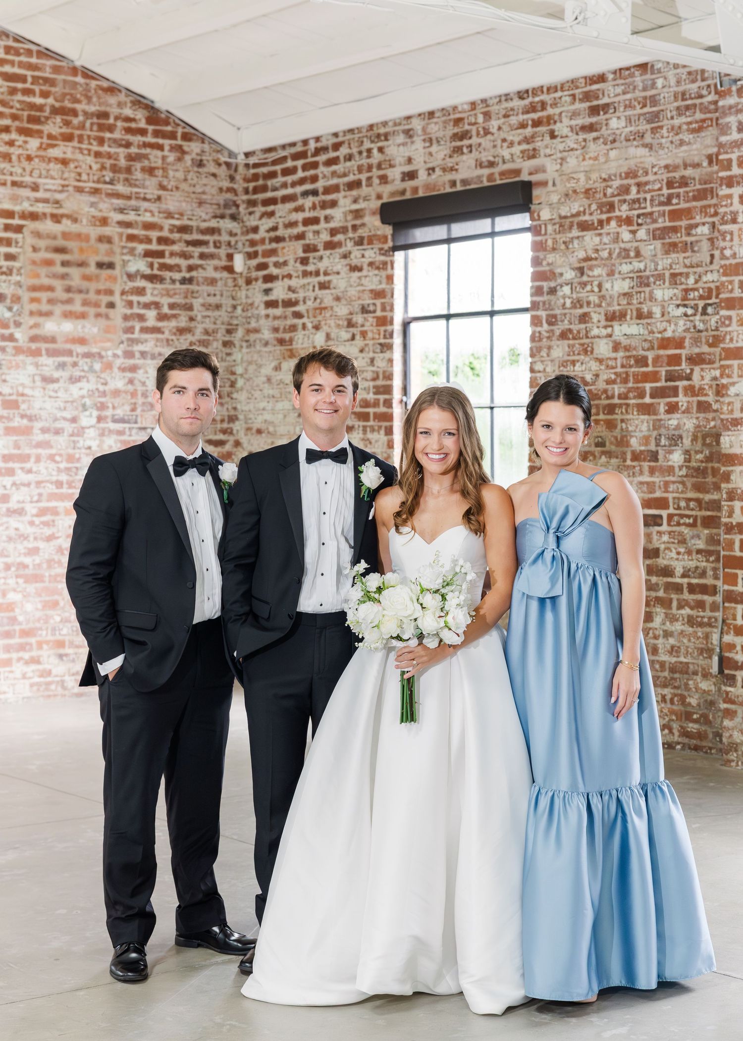 Wedding party of four posing in brick venue with bride in white gown and bridesmaid in blue dress.